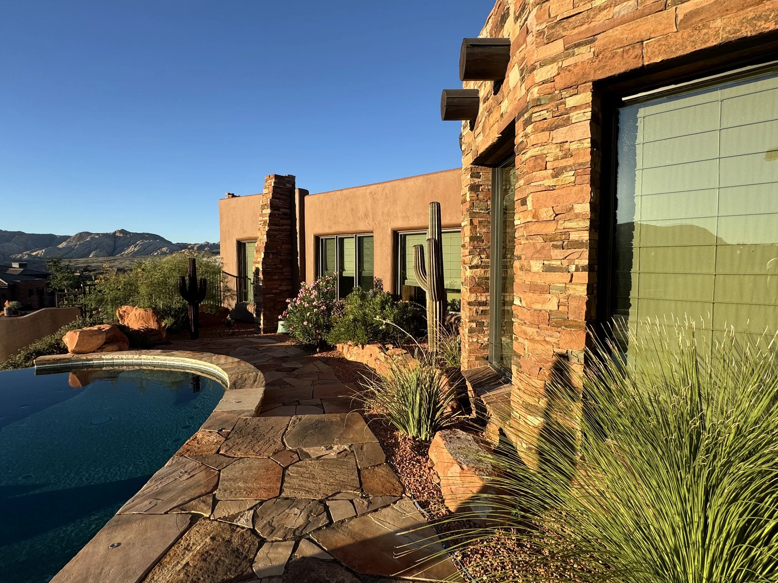 A desert-style backyard with a kidney-shaped pool bordered by natural stone pavers, set against a modern house with stone and stucco exterior walls, cacti, lush desert plants, and a mountain landscape in the background under a clear blue sky.