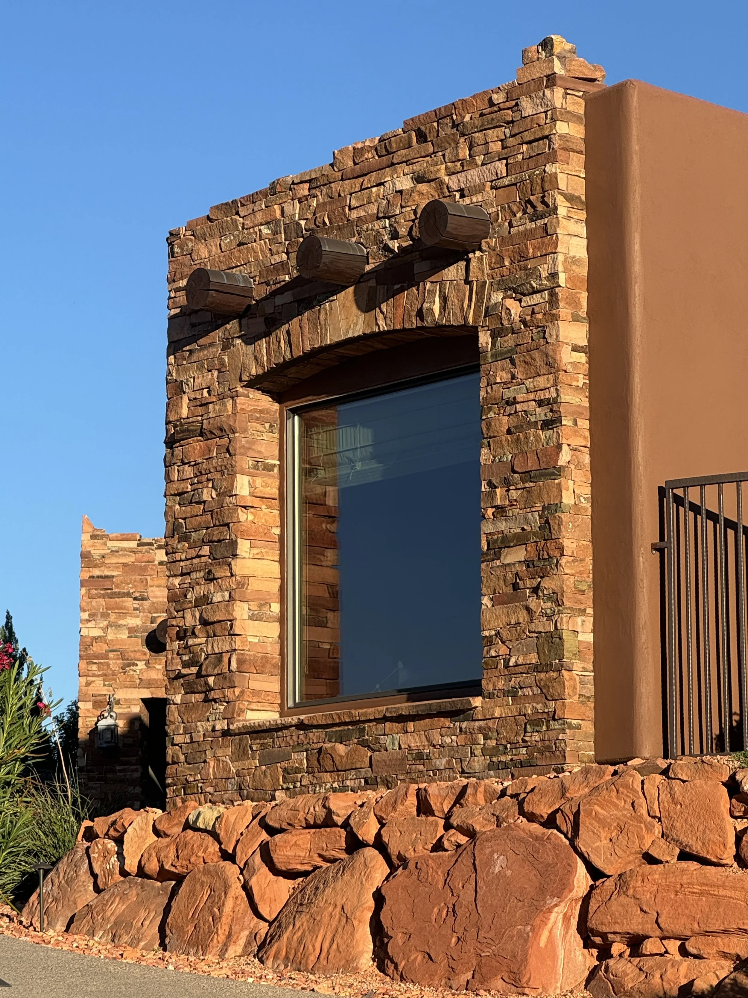Close-up of a building with a brick and adobe exterior, a large window, and a stone landscaping base under a clear blue sky.