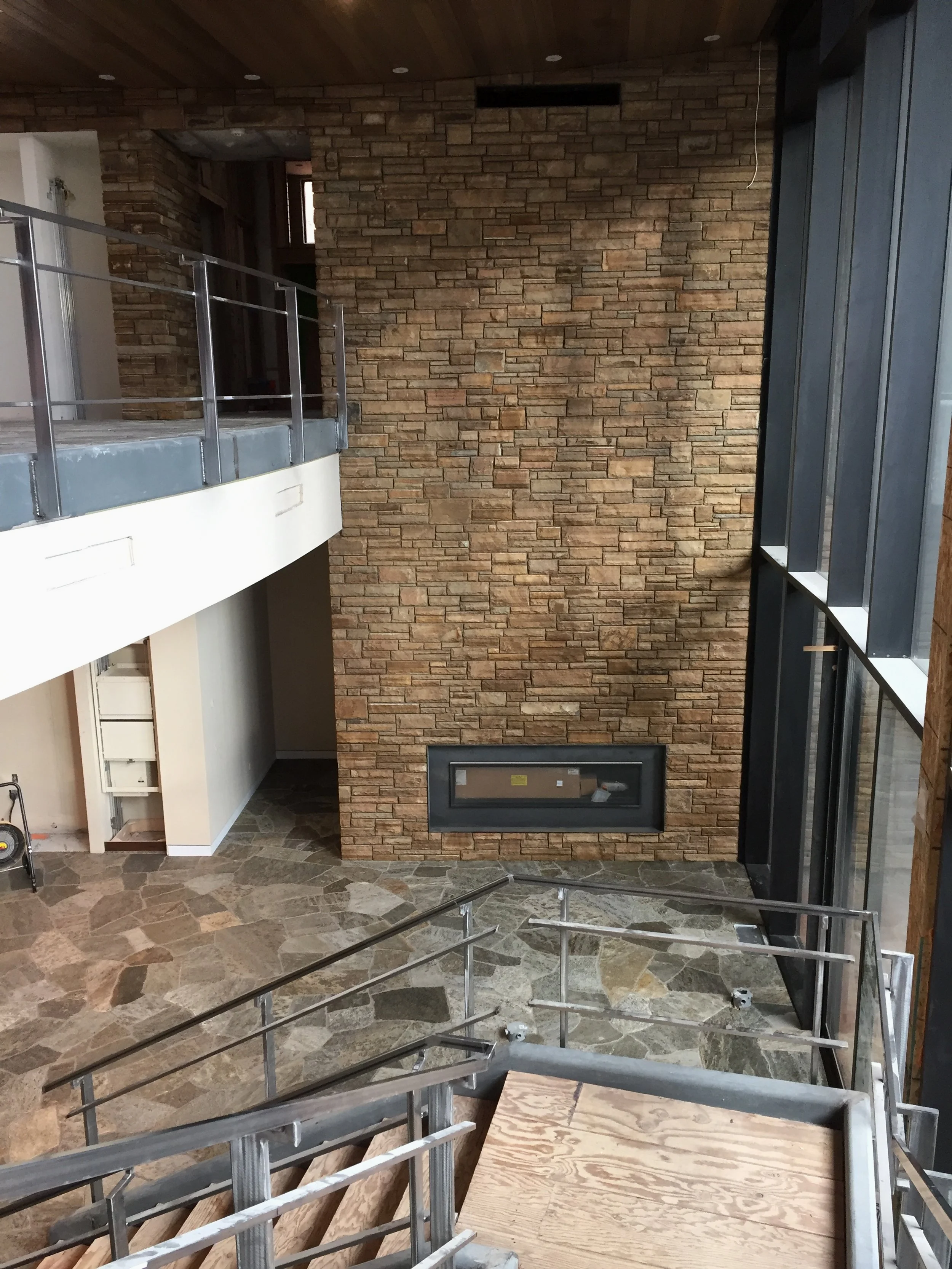 Interior of a building with a stone accent wall, large windows, and a fireplace opening, with construction materials and scaffolding present.