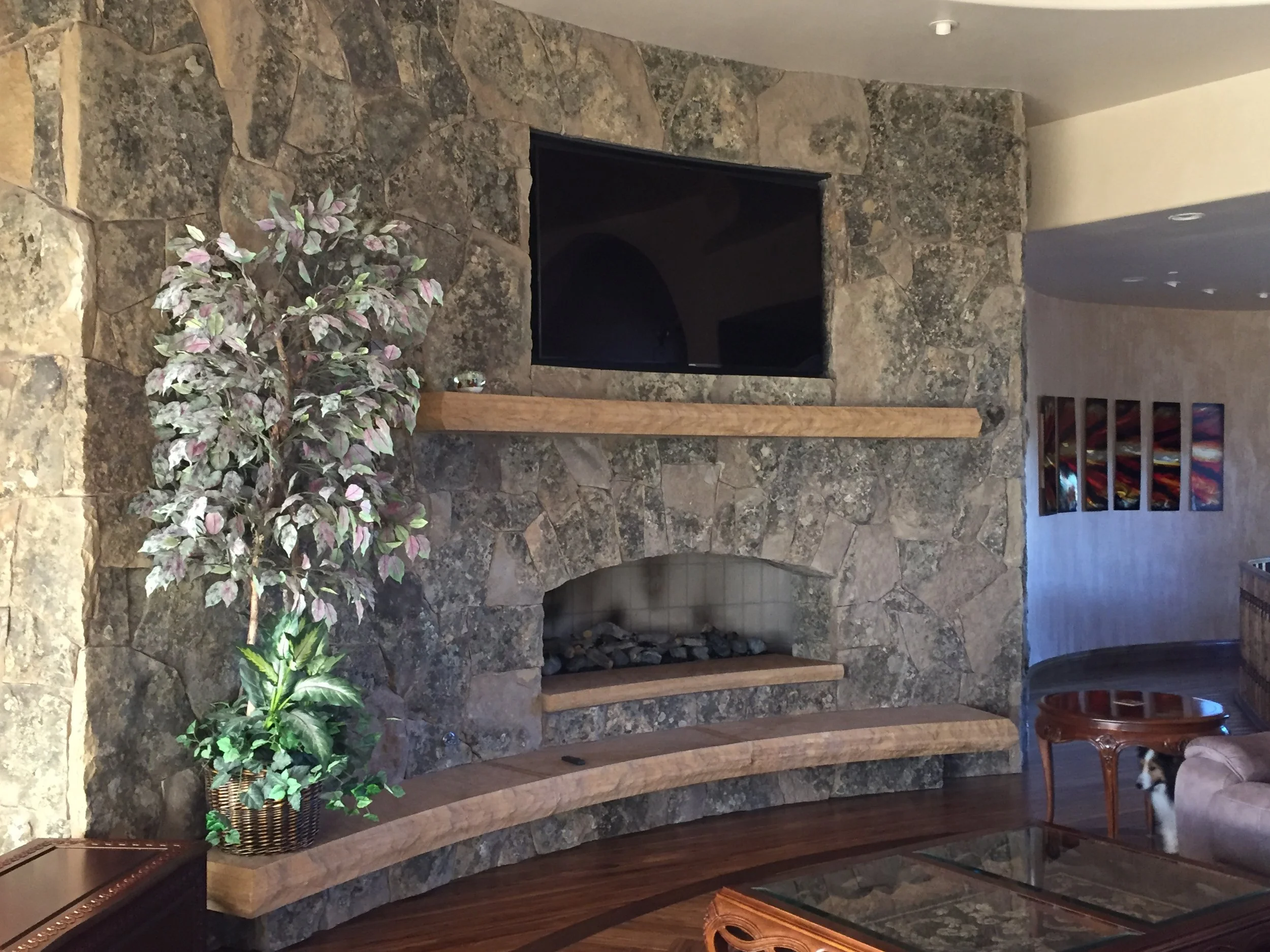 A living room with a large stone fireplace, a flat-screen TV above the fireplace, a wooden mantel, a potted plant on the left side, a small dog on the right side under a round wooden table, colorful artwork on the wall, and a glass-topped coffee table.
