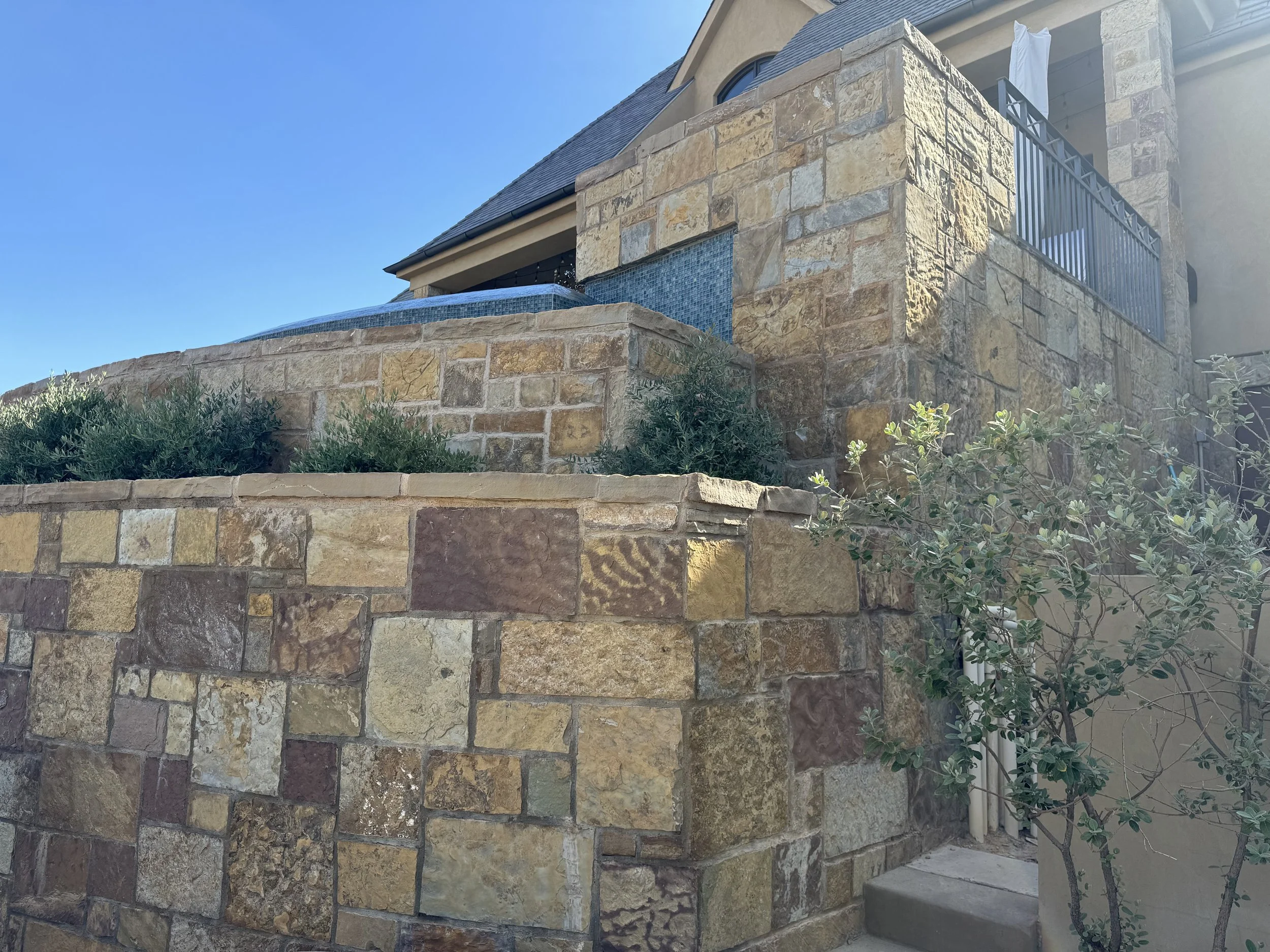 Stone retaining wall with plants and shrubs, part of a house with a balcony and laundry hanging out to dry, clear sky in the background.