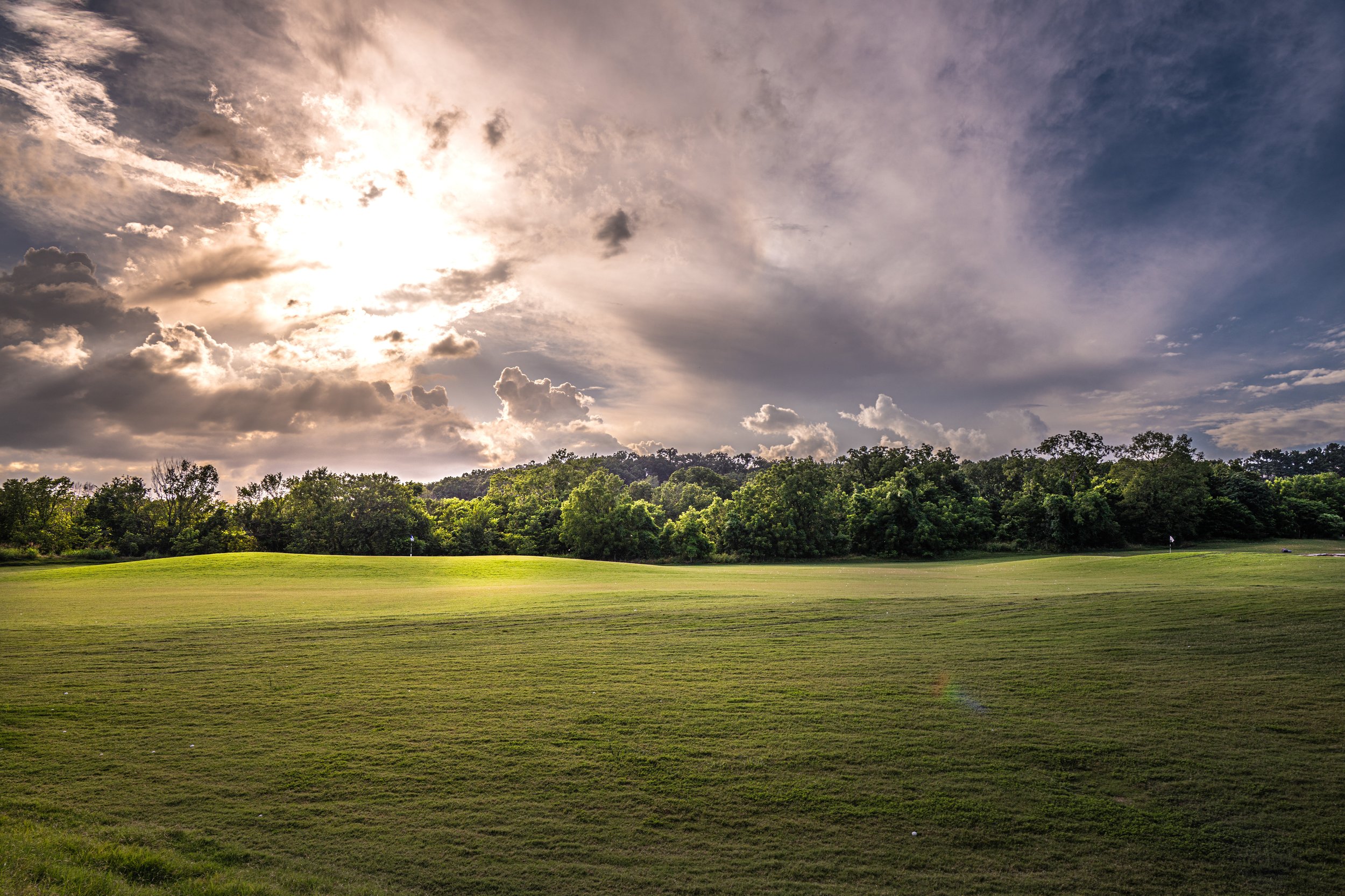 Wide grassy field with tree line under a dramatic sky with sunlight and clouds in Bentonville, Arkansas.