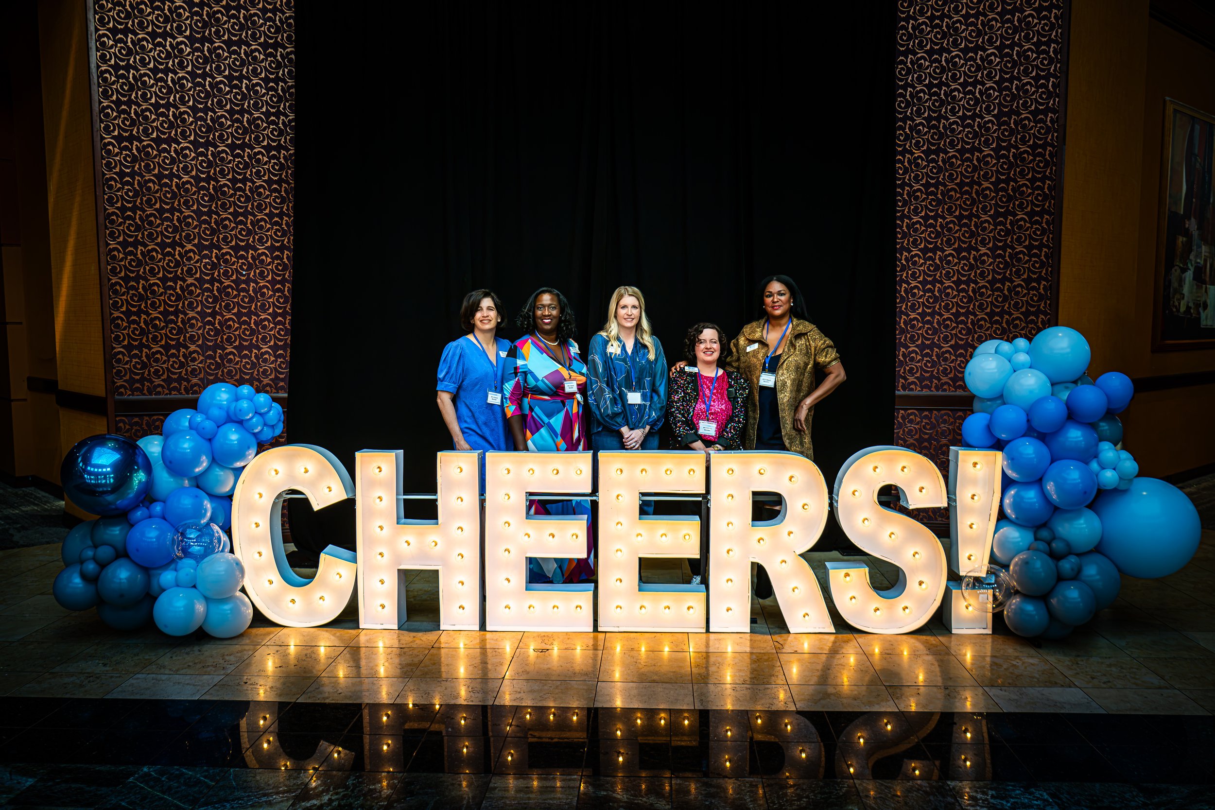 Premier Chef Northwest Arkansas 2026 event organizers pose behind illuminated CHEERS letters and blue balloons at the Rogers Convention Center.