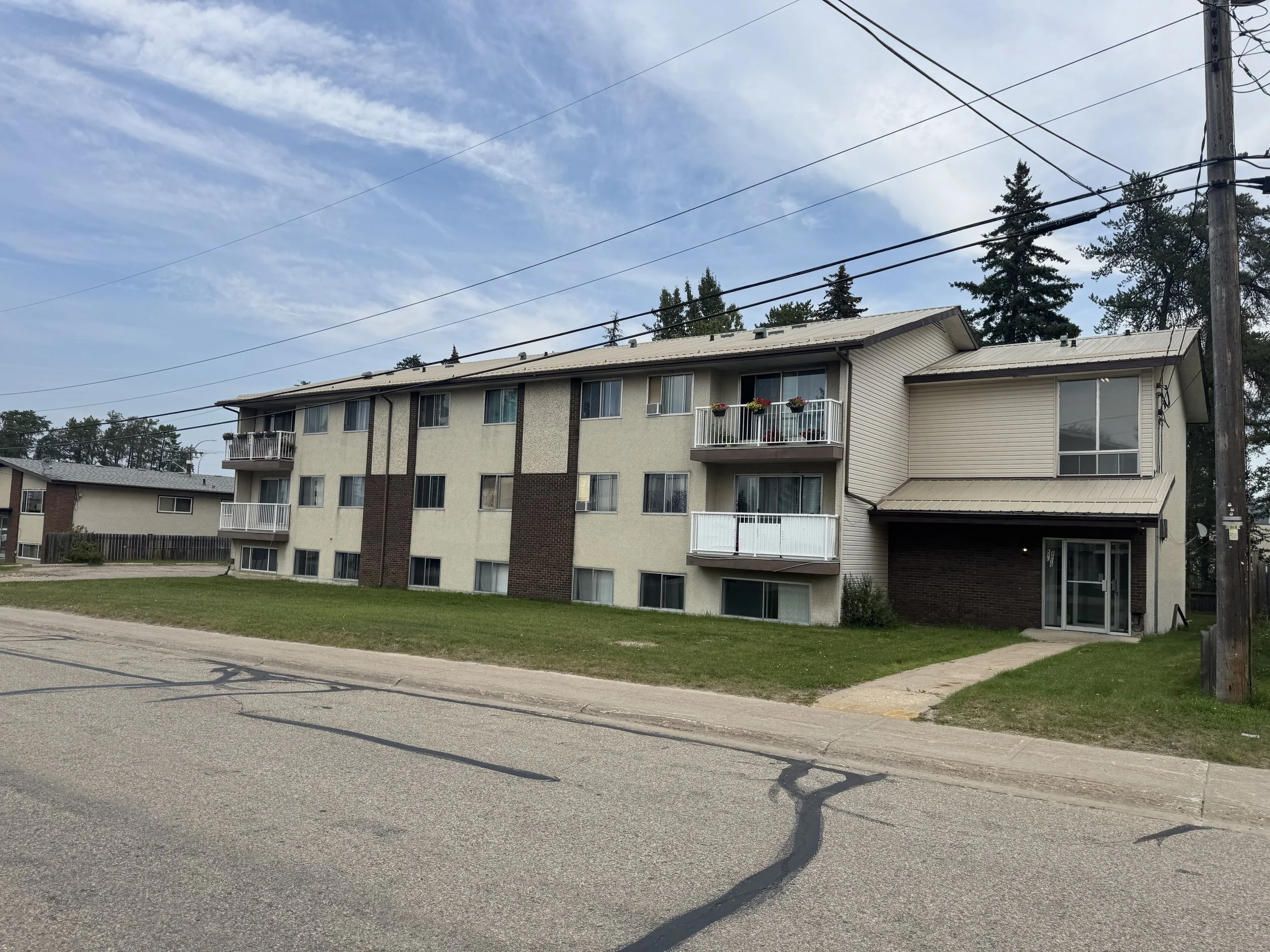 Three-story residential apartment building with beige siding and brown brick accents, balconies with white railings, and a manicured lawn in front, under a partly cloudy sky.