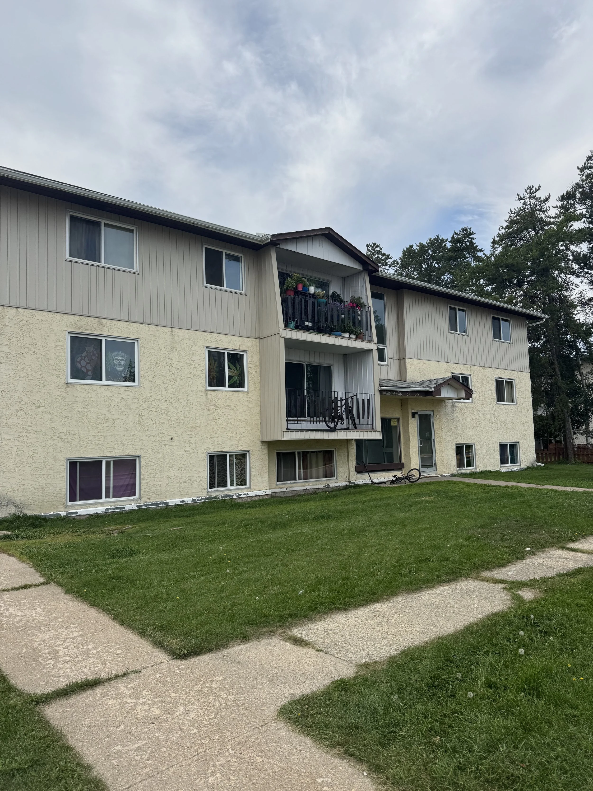 View of a multi-story apartment building with balconies, some with bikes and flower pots, and a grassy lawn with concrete pathways in the foreground.