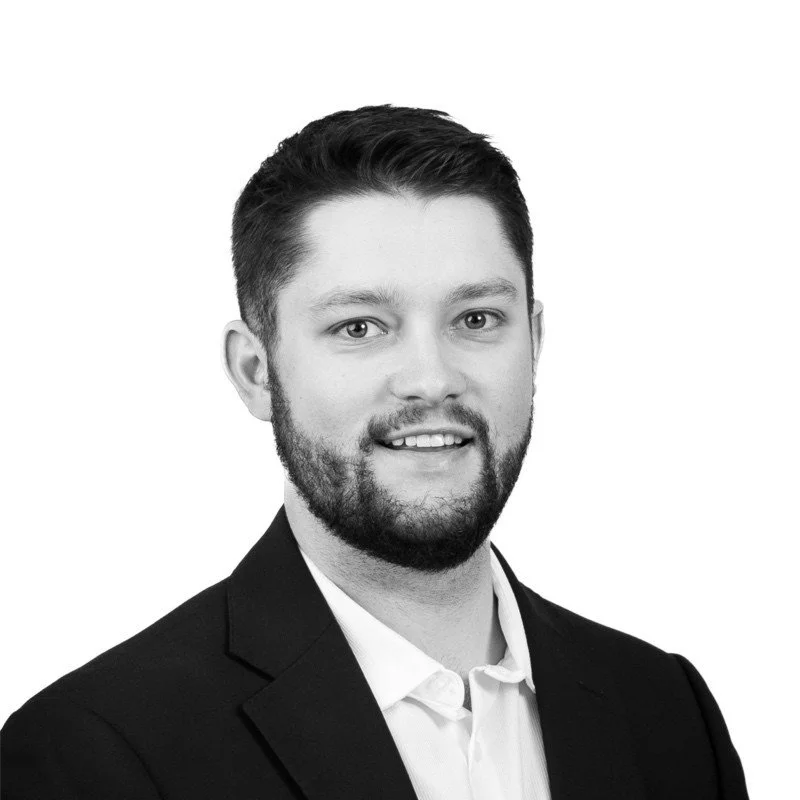 Black and white headshot of a young man with short hair, beard, wearing a suit jacket and collared shirt, smiling slightly