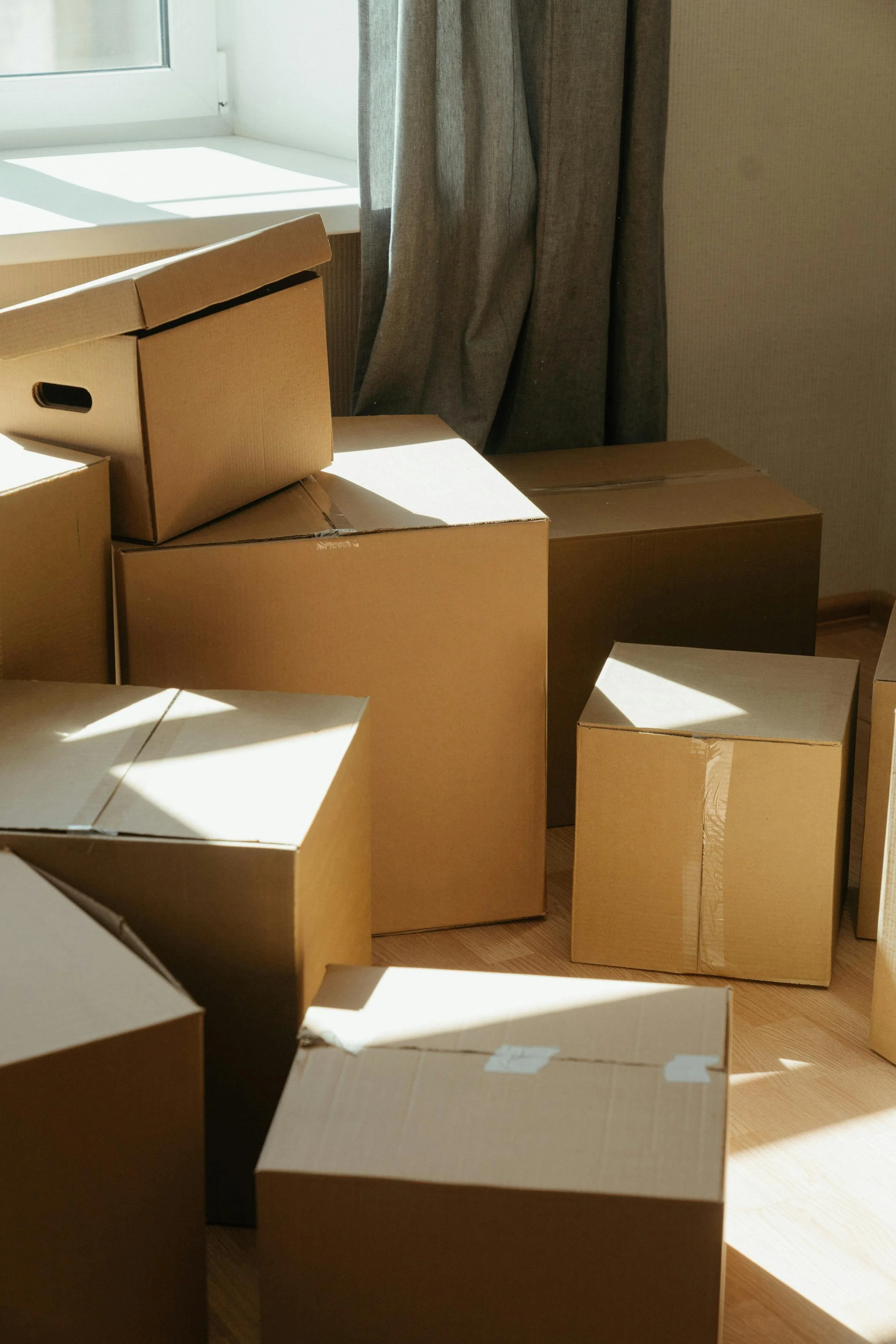 Several cardboard boxes of various sizes placed on a wooden floor near a window with sunlight streaming in, partially covered by a curtain.