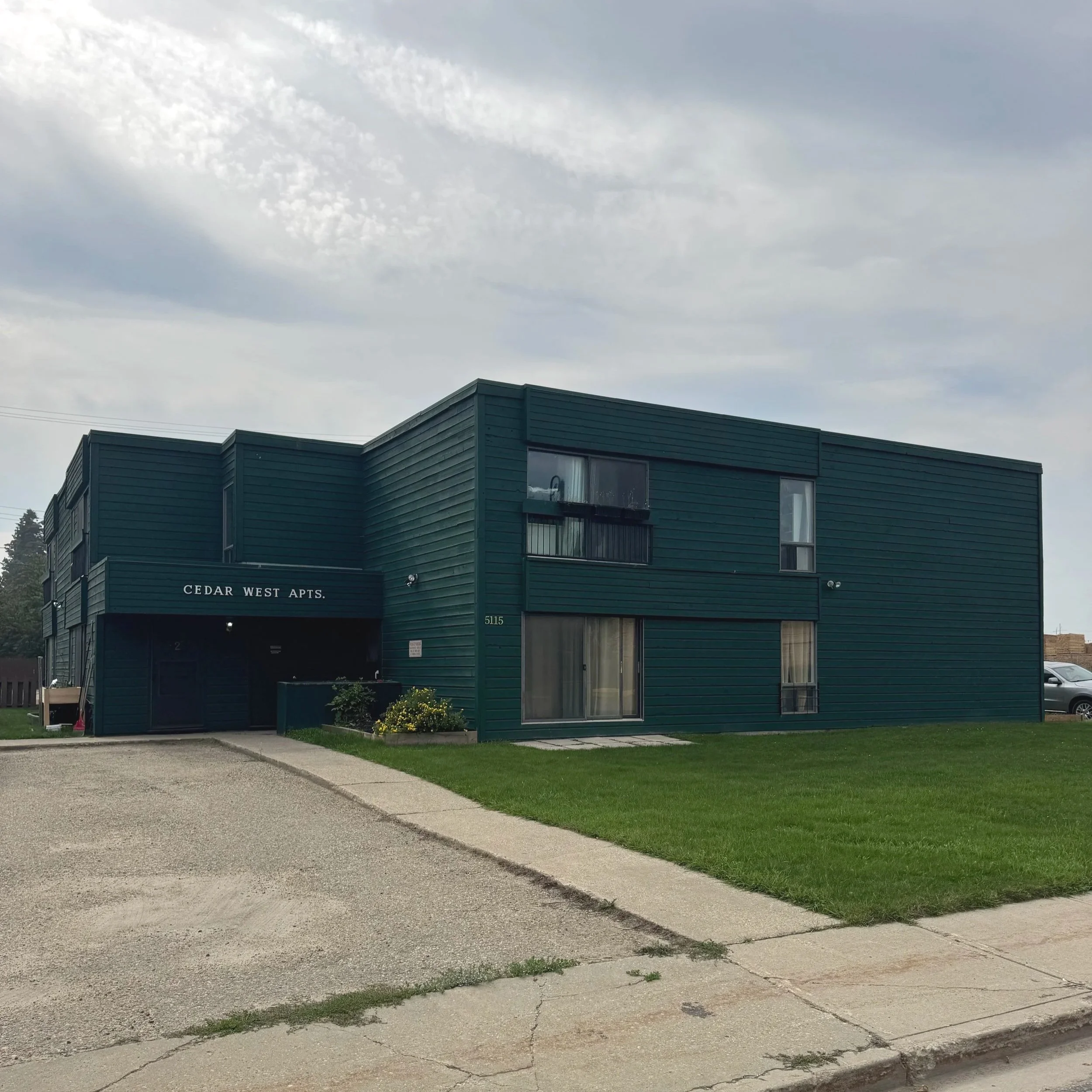 A dark green multi-unit apartment building with a sign that reads 'Cedar West Apts.' in white lettering. The building has several windows, some with curtains, and a small parking lot with parked cars nearby.