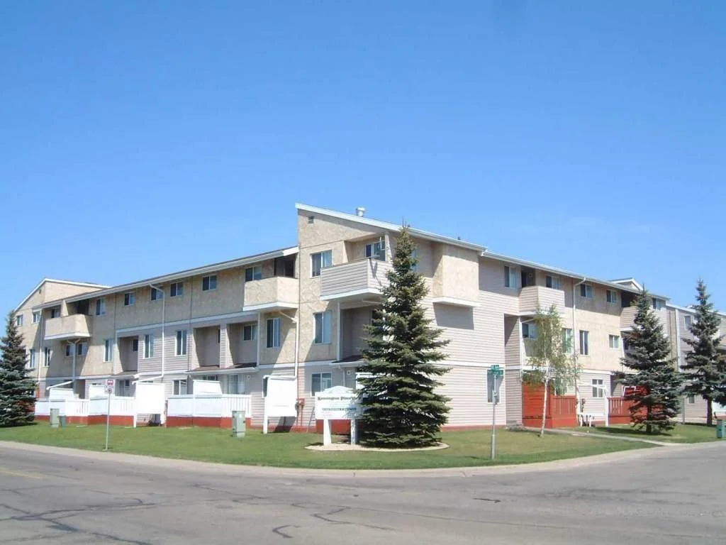 A multi-story residential apartment building with beige and light gray exterior, surrounded by a green lawn, pine trees, and a clear blue sky.