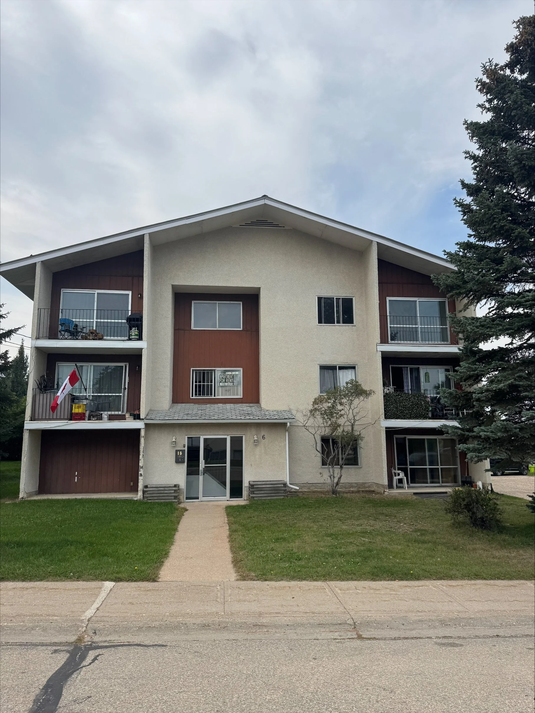 Four-story apartment building with balconies, entrance door, and a small lawn in front, situated in a residential area with trees and a sidewalk.
