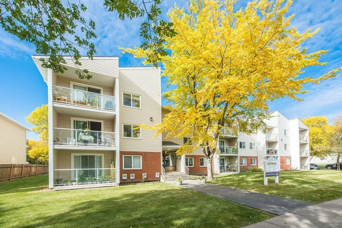 Three-story apartment building with balconies, surrounded by green grass and trees, under a partly cloudy sky.