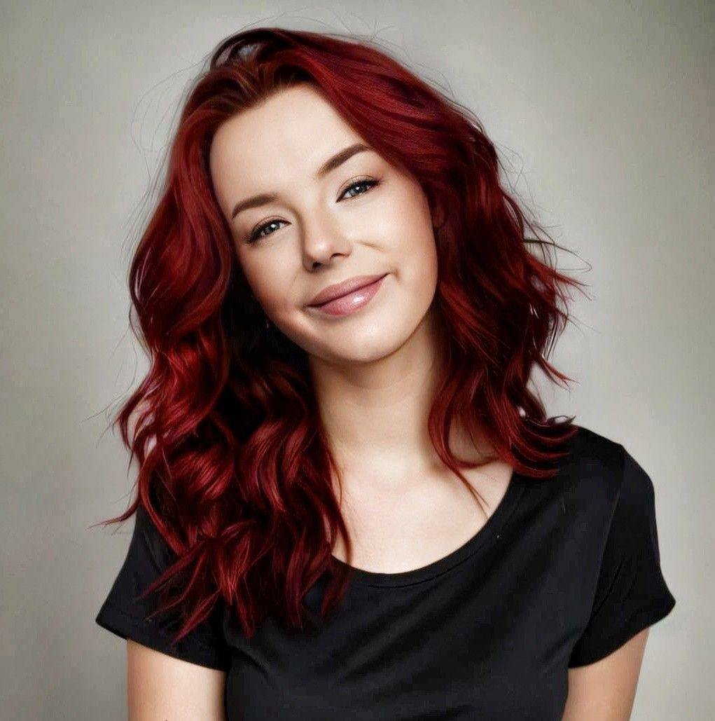 A woman with shoulder-length wavy red hair, smiling slightly, wearing a black t-shirt against a light-colored background.