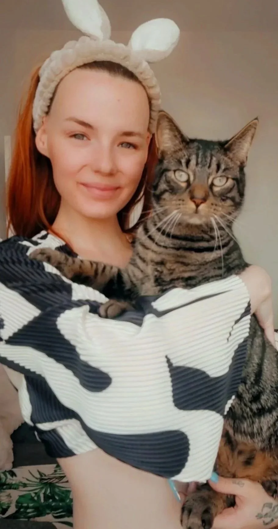 A woman with red hair wearing a bunny ear headband, holding a large tabby cat with striped fur and green eyes, against a plain wall background.