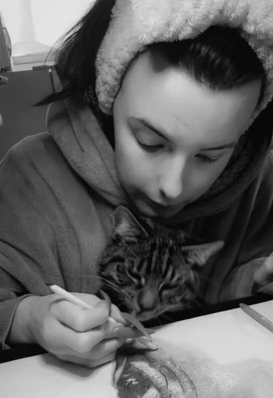 A woman in a hooded sweatshirt with a fuzzy headband is drawing a tiger on paper, holding a pencil, with a cat resting on her chest.