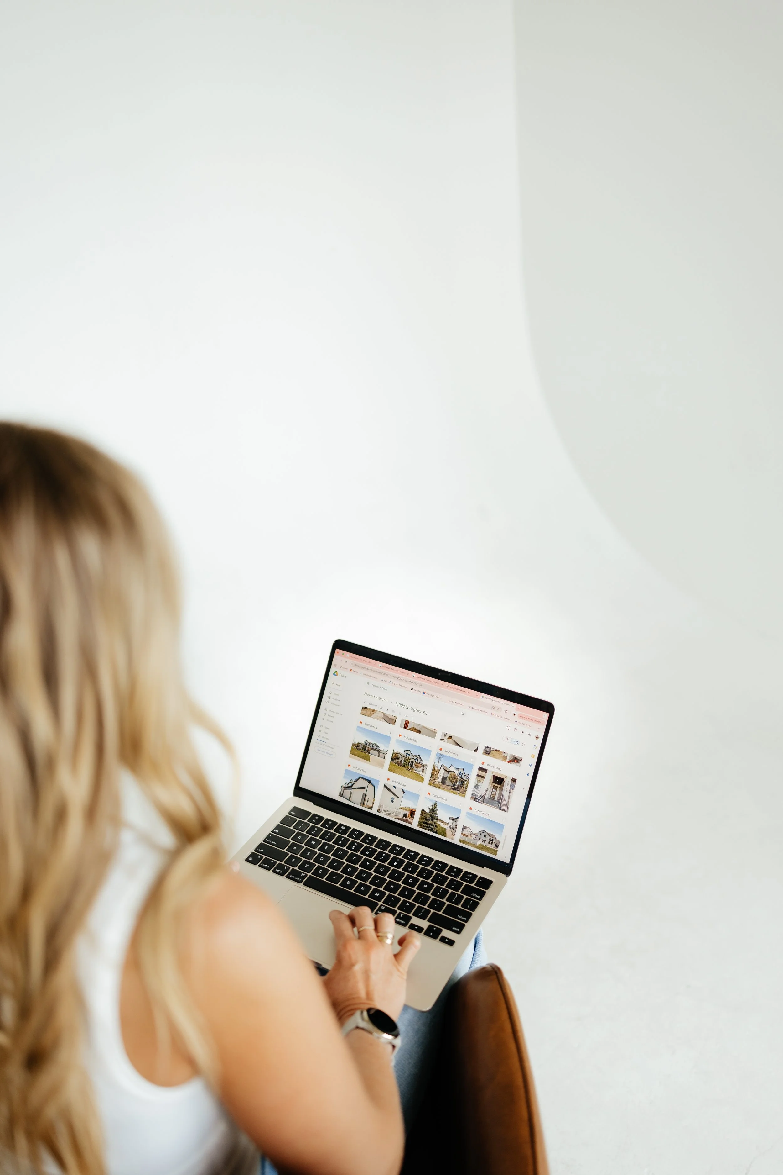 A woman with long blonde hair sitting in a chair, browsing real estate listings on a laptop.