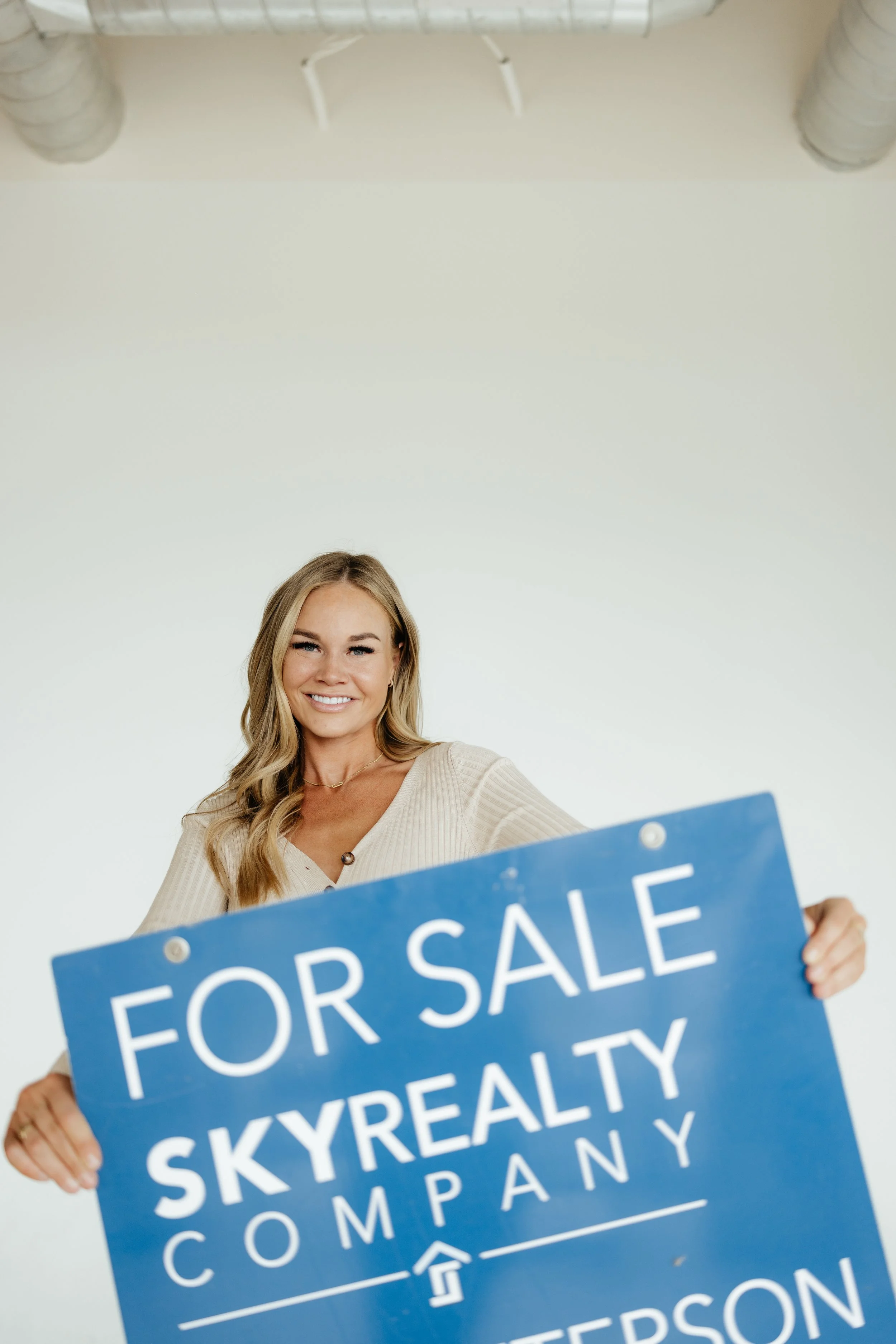 A smiling woman holding a large blue sign that reads 'For Sale SkyRealty Company' in an empty room with white walls and ductwork overhead.