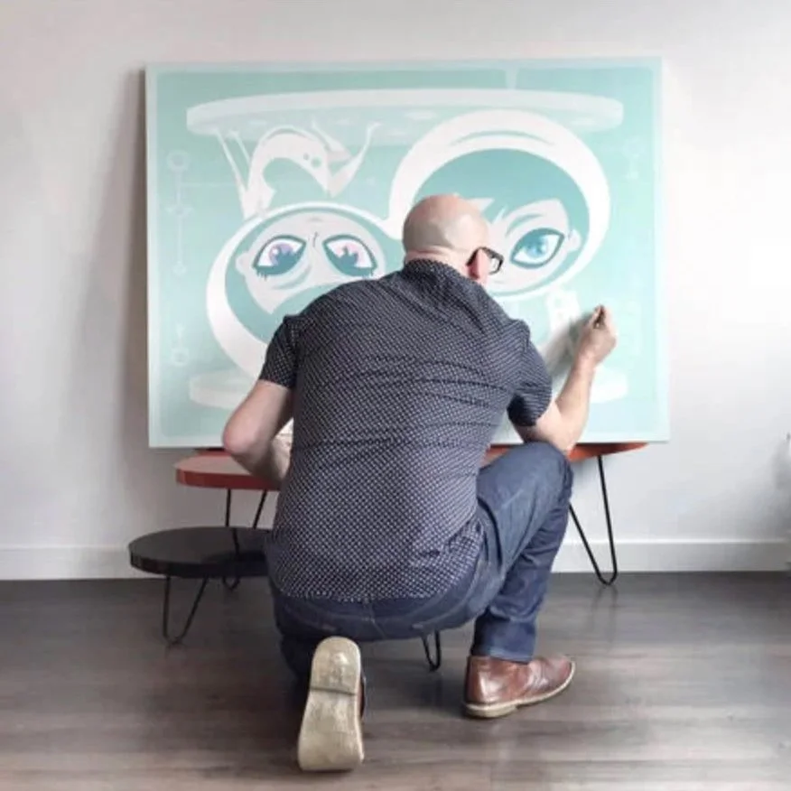 An artist kneeling on one knee, working on a large, light blue abstract painting with a cartoonish face, glasses, and eyes, against a plain white wall in a studio or gallery.