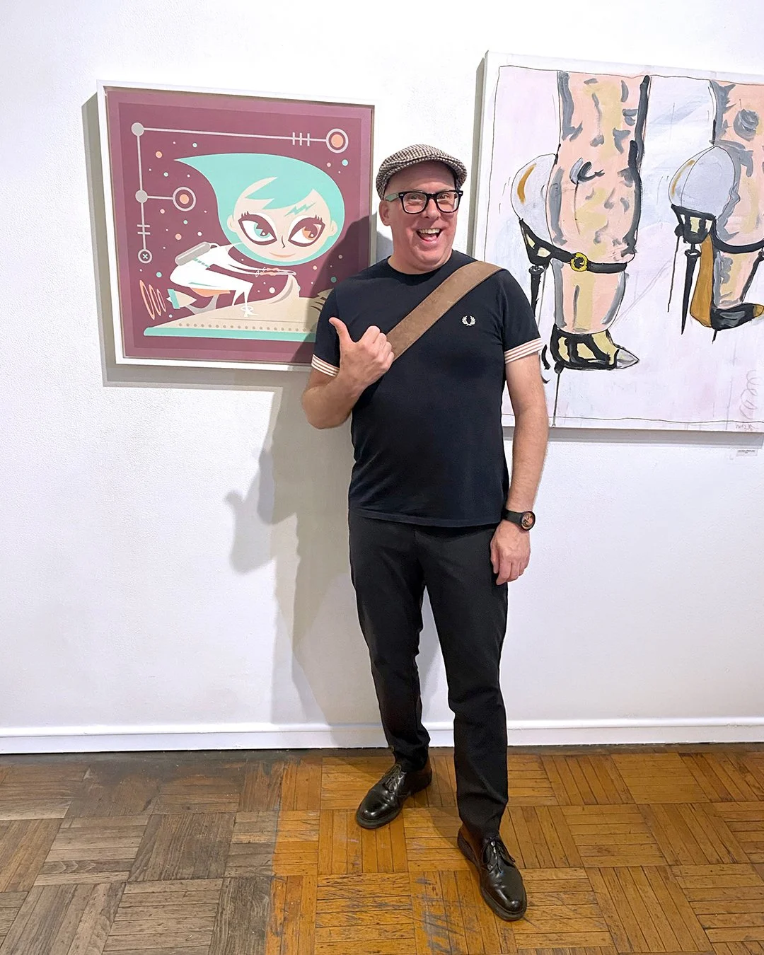 Artist Mark Atomos Pilon wearing a black Fred Perry t-shirt, and black pants standing in front of his colourful artwork. He is smiling and pointing at the artwork on his left, he's at the La Luz de Jesus Gallery opening of their 40th Anniversary.