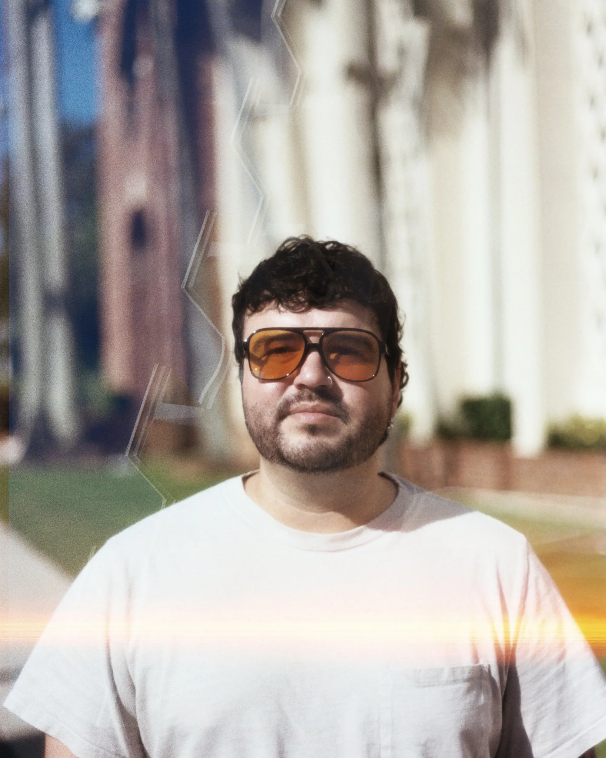 A person with short dark hair and glasses wearing a white t-shirt, standing outdoors in front of a blurred urban background.