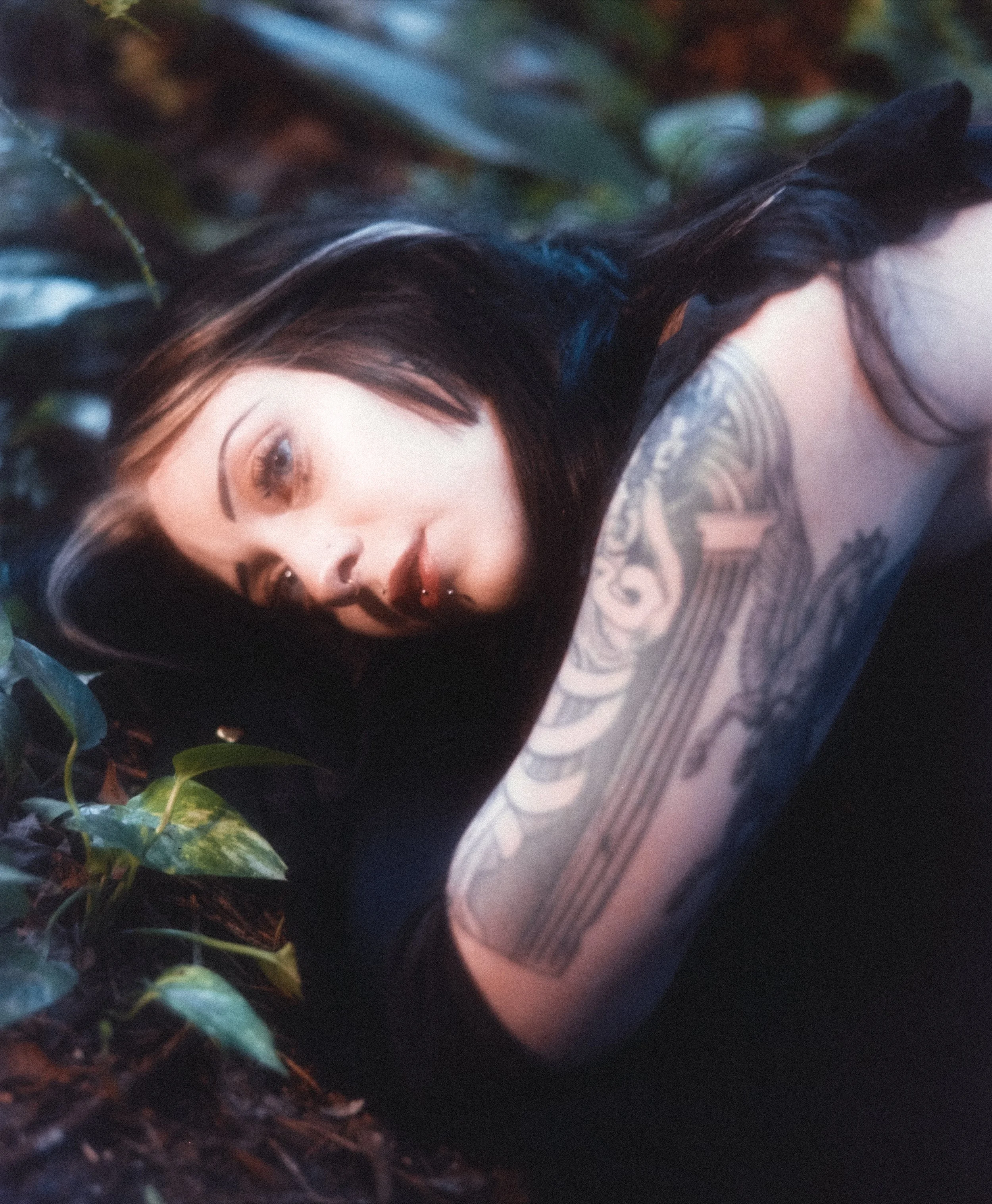 A woman with black hair and tattoos lying among green leaves with a serious expression.