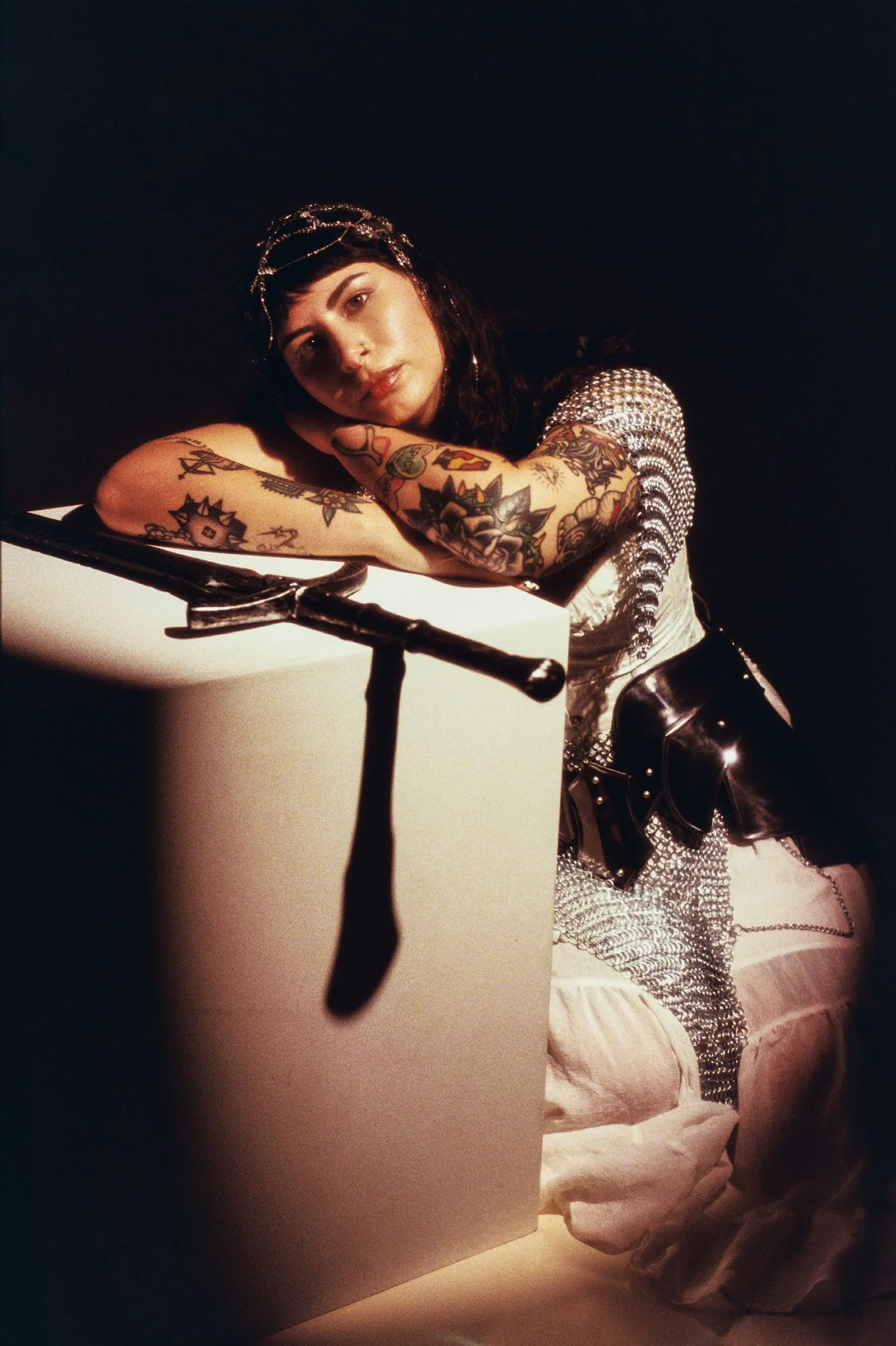 A woman with dark hair, tattoos on her arms, wearing a silver headpiece and a silver mesh top, leaning on a white surface in a dark room.