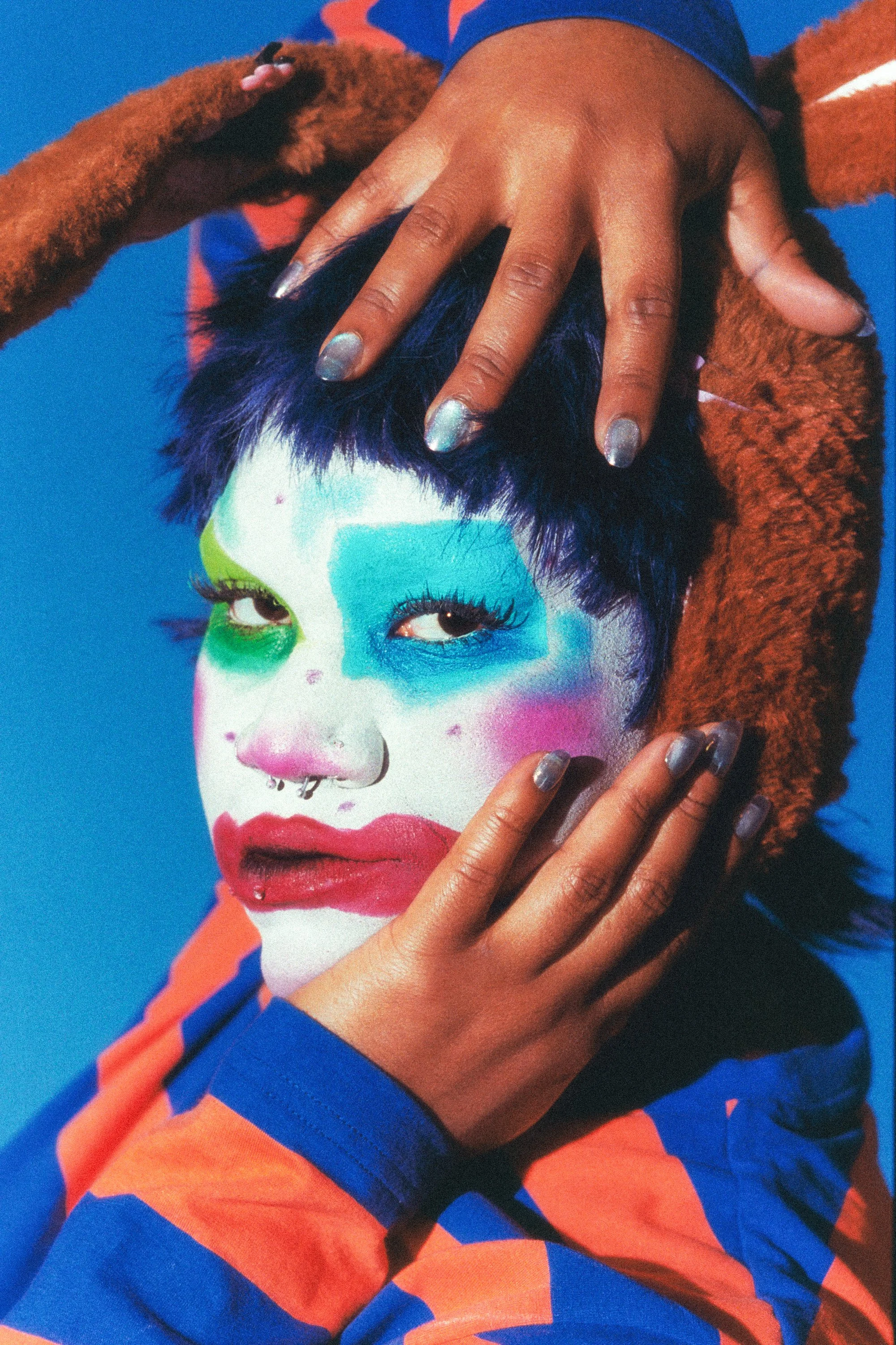 A person with vibrant makeup and colorful clothing, posing with hands around their head, against a bright blue background.