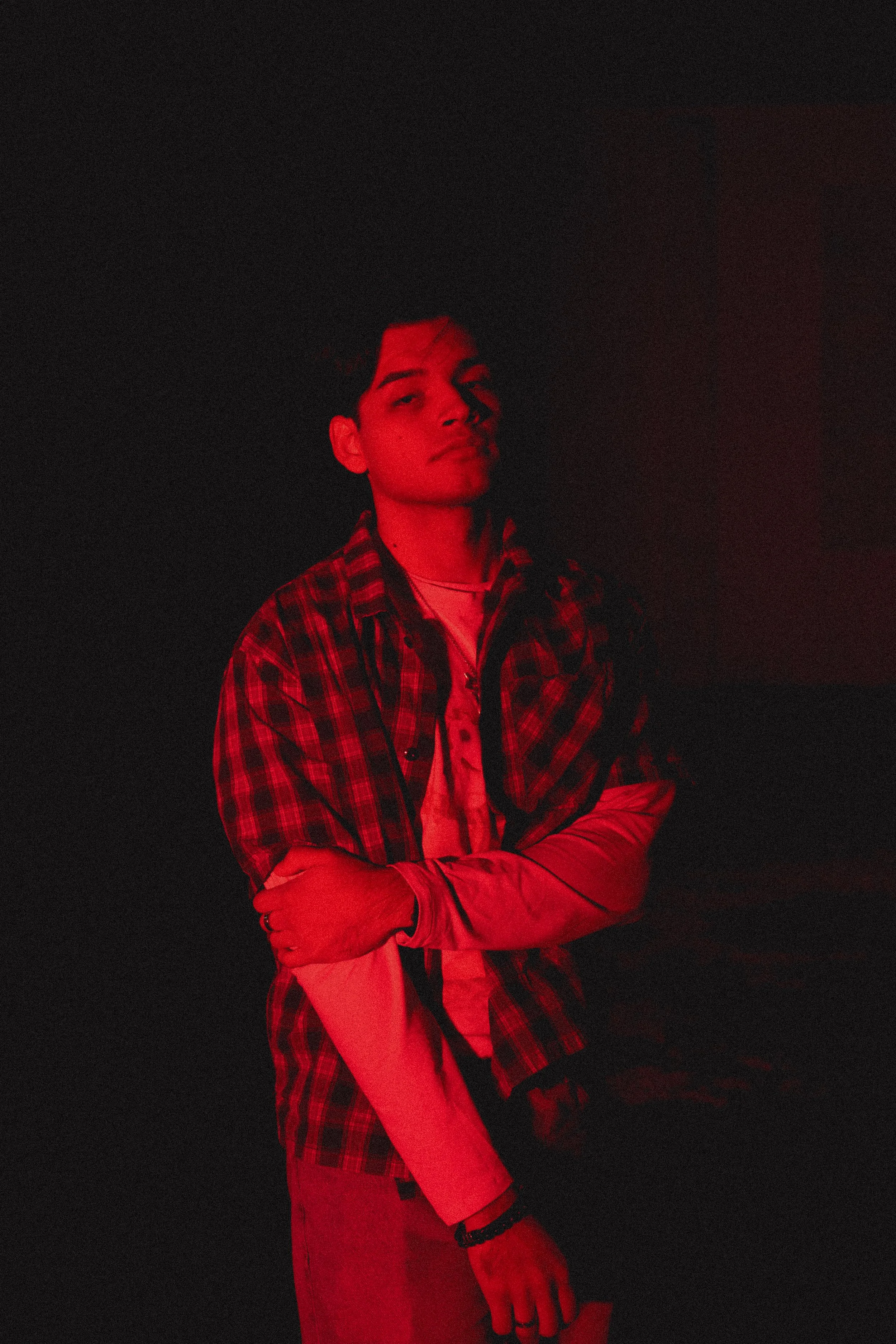 A young man with arms crossed, standing in front of a dark background, illuminated with red light.