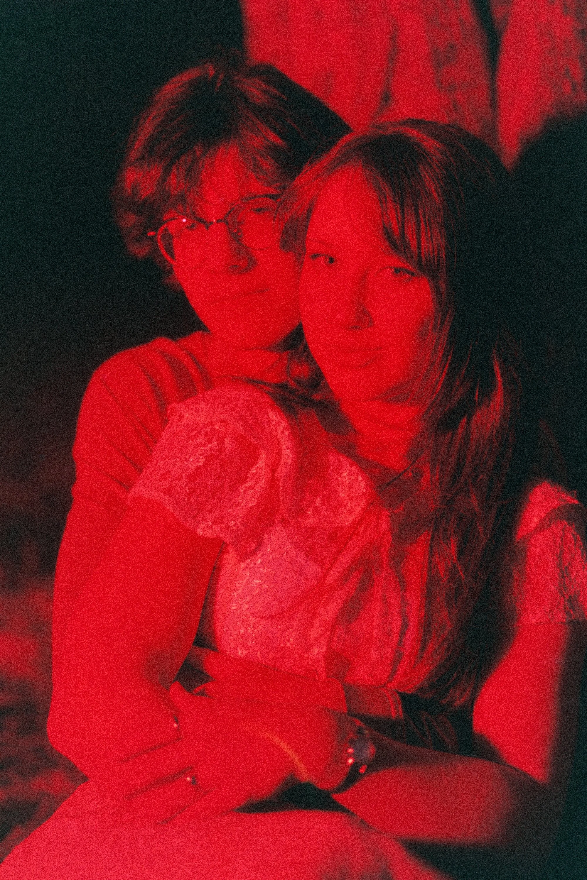 A woman in lace clothing with long hair is taking a selfie in a dark room with red lighting, with another woman's face partially visible beside her.