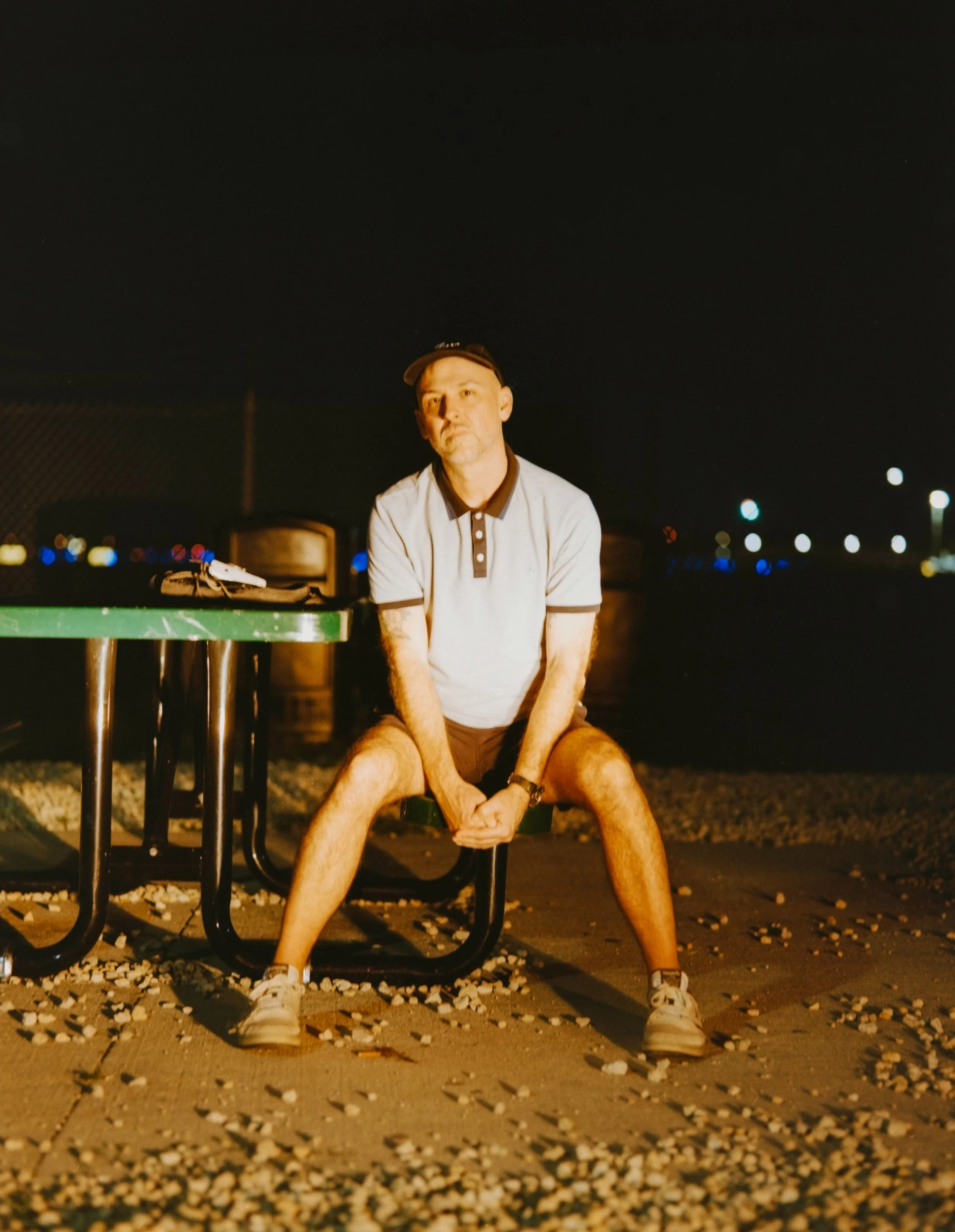 A man with a beard wearing a polo shirt and shorts squatting with his arms crossed in front of him, at night, with a dark sky and city lights in the background, and a green-painted table in front of him with some objects on top.