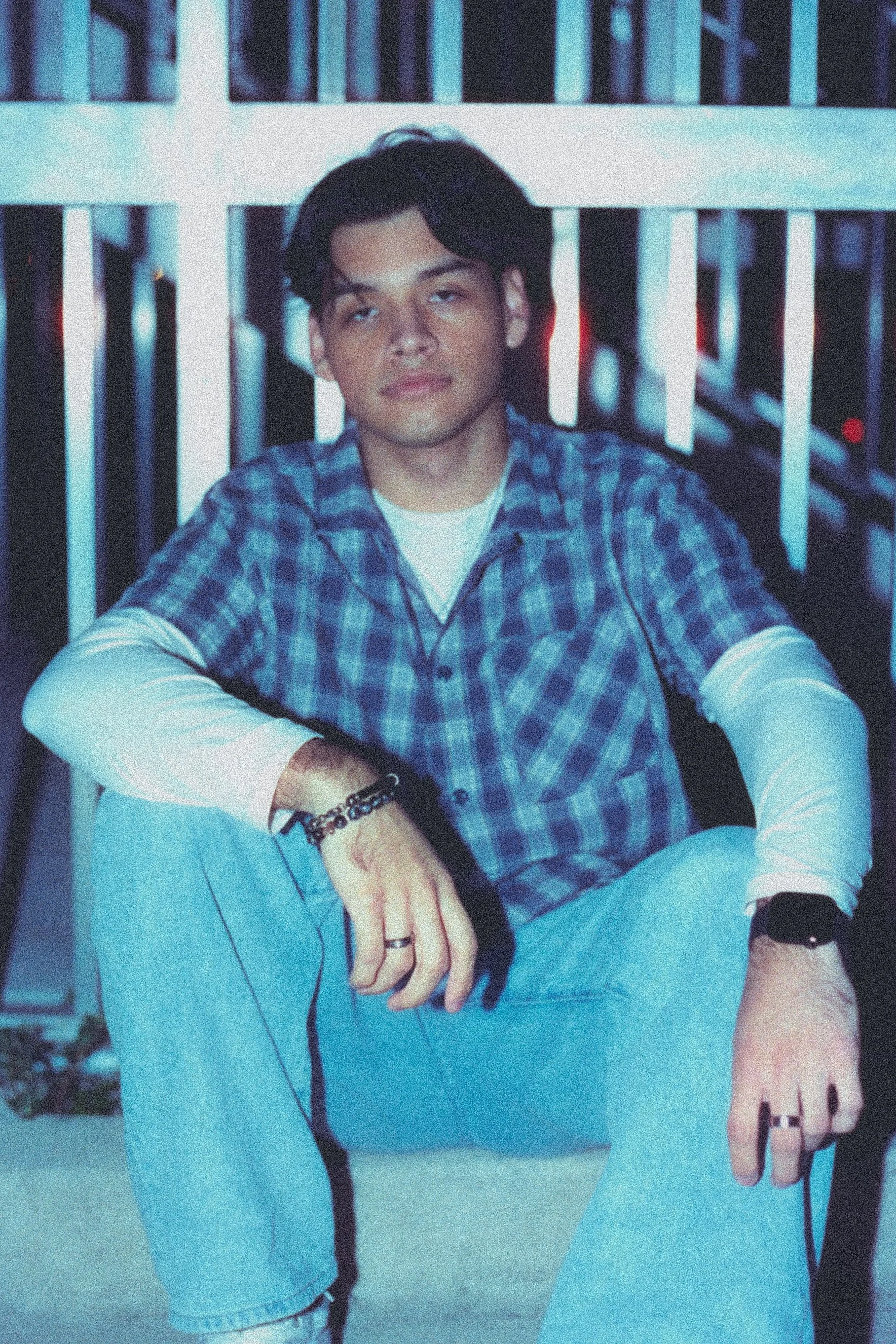 A young man with dark wavy hair wearing a blue plaid shirt and white long sleeve shirt, sitting against a background of a white railing with dark shadows, looking slightly cross-eyed with a relaxed expression.