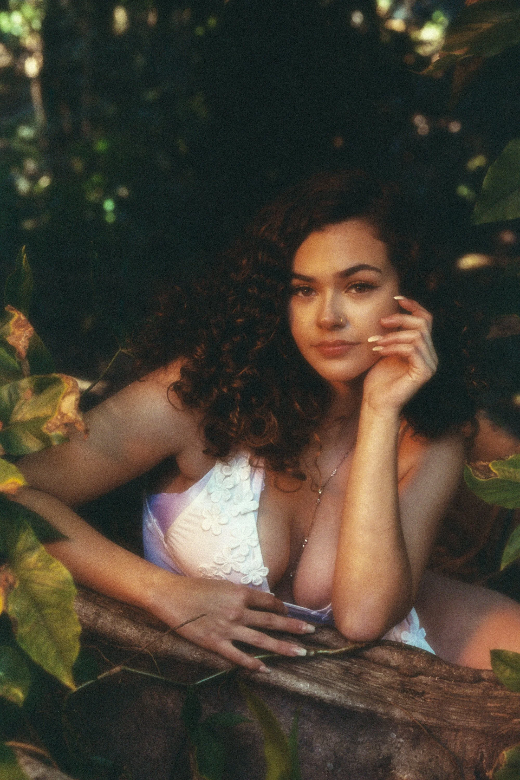 A woman with curly brown hair, wearing a white dress with floral embroidery, is resting her chin on her hand while lying on a large tree branch surrounded by green leaves.