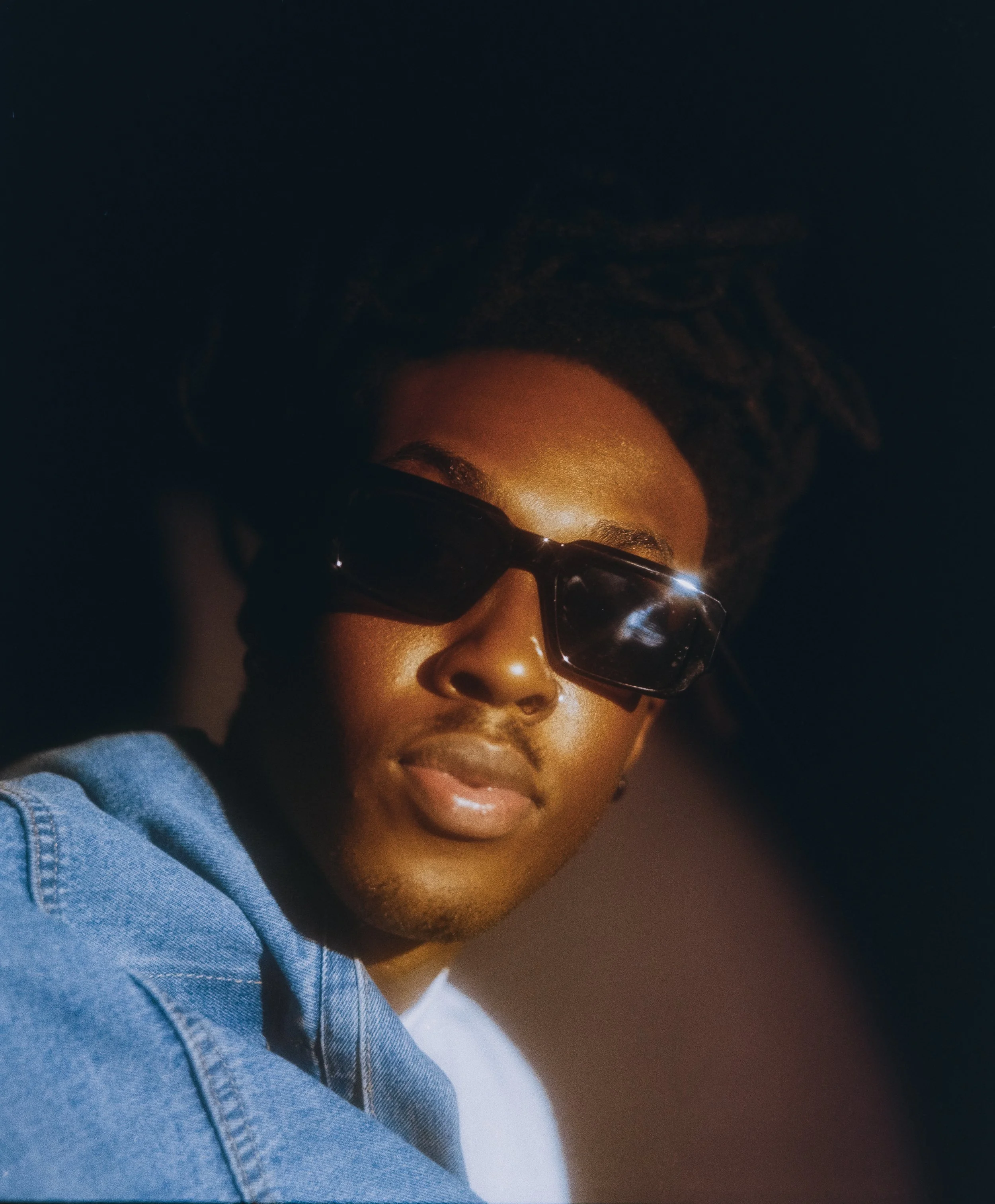 Close-up of a person's face with curly hair, wearing glasses, and dressed in a denim jacket over a white shirt, against a dark background.