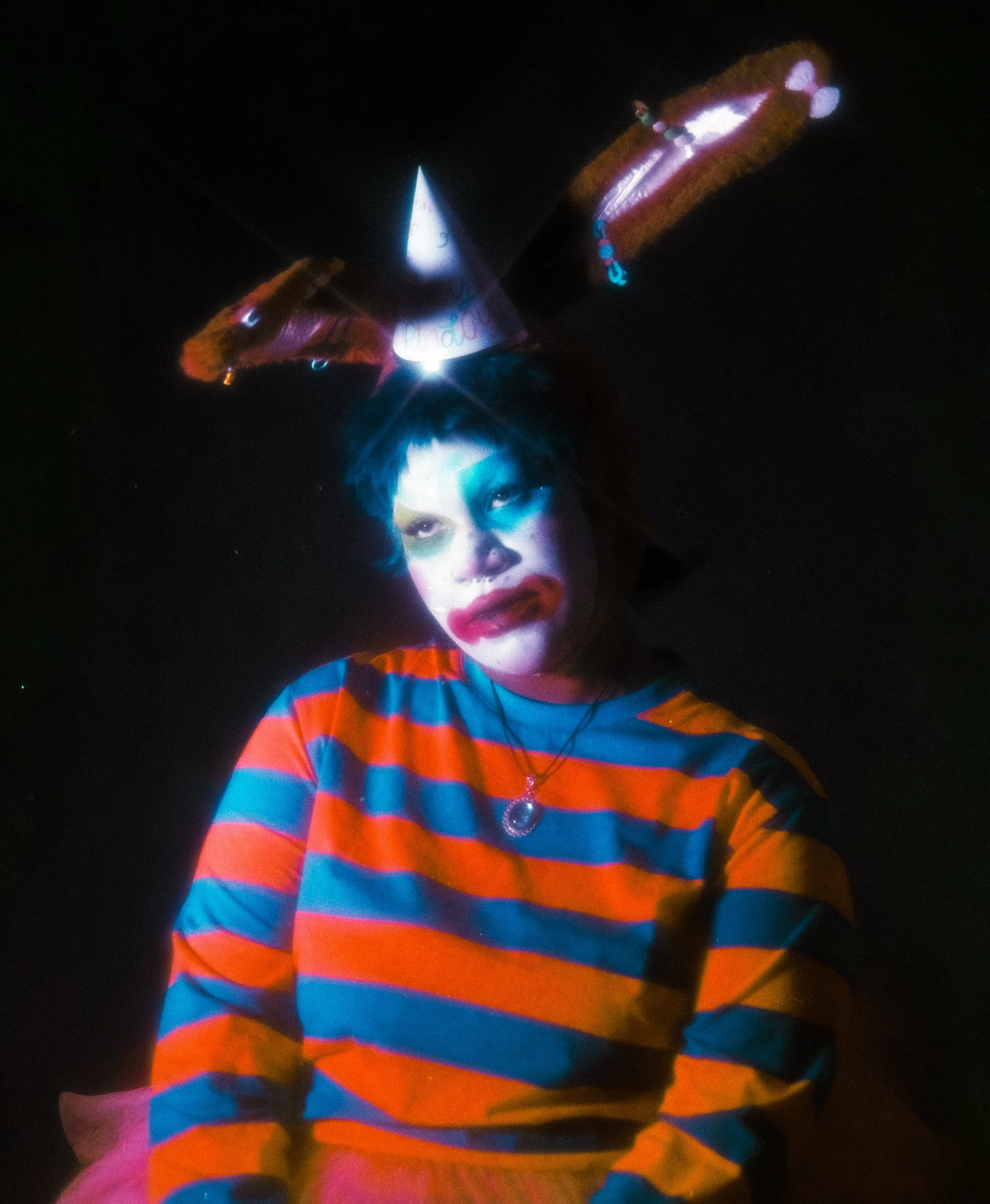 Person with colorful makeup and striped shirt in a dark setting with neon or glow-in-the-dark elements.