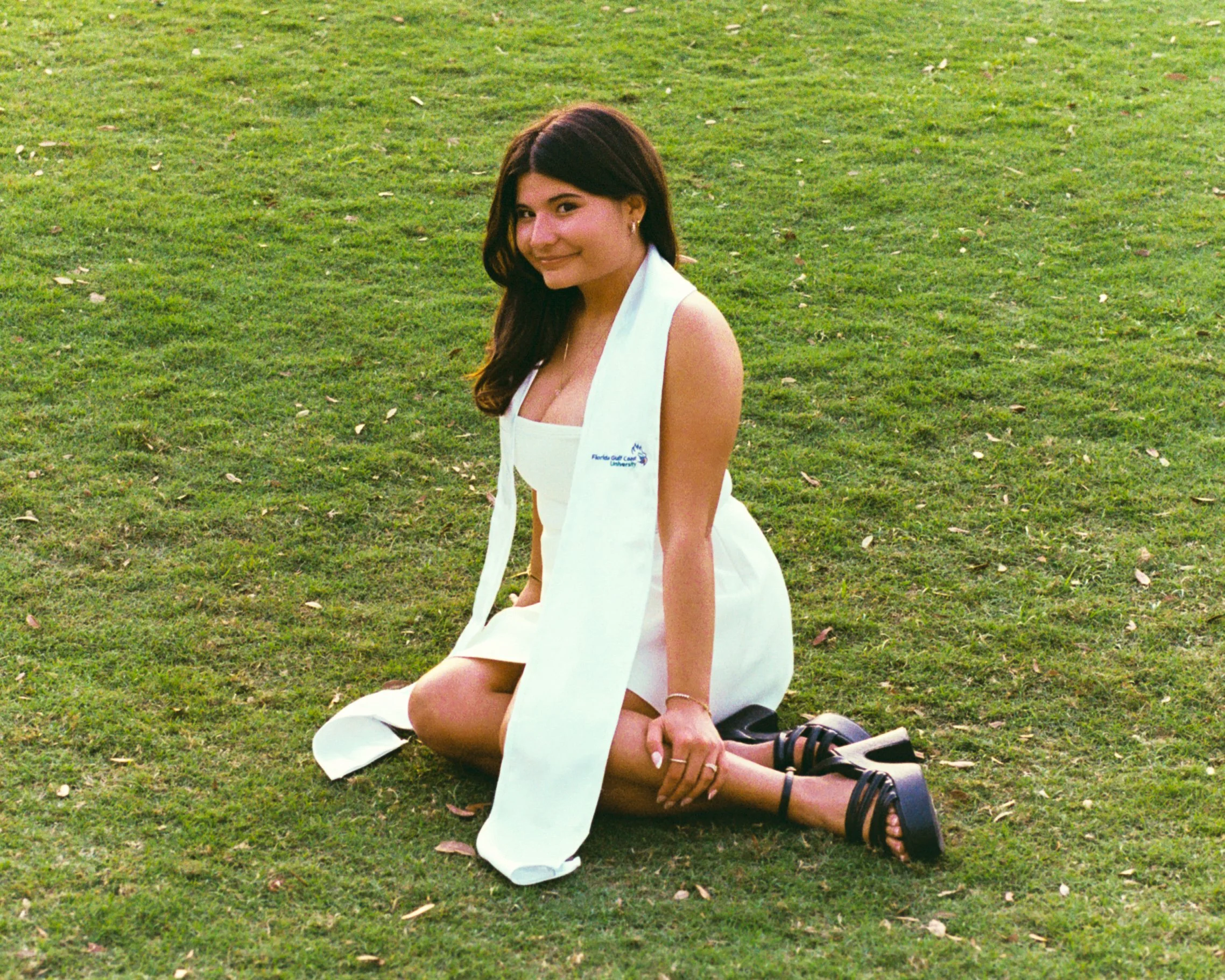A young woman with long dark hair, wearing a white dress, graduation outfit, and black strappy sandals, sits on the grass with her legs bent to the side, smiling at the camera, in an outdoor setting.