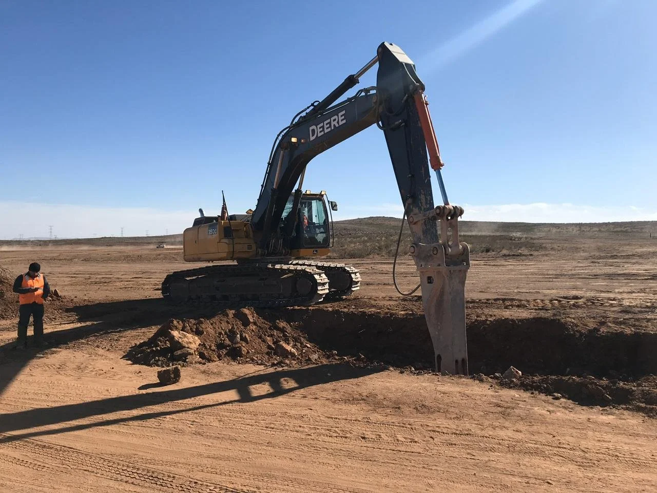 Excavadora John Deere operada por Aude Energía en el proyecto BNB Villahumada Solar I, realizando excavación de zanjas para infraestructura en parque solar, con terreno desértico al fondo.