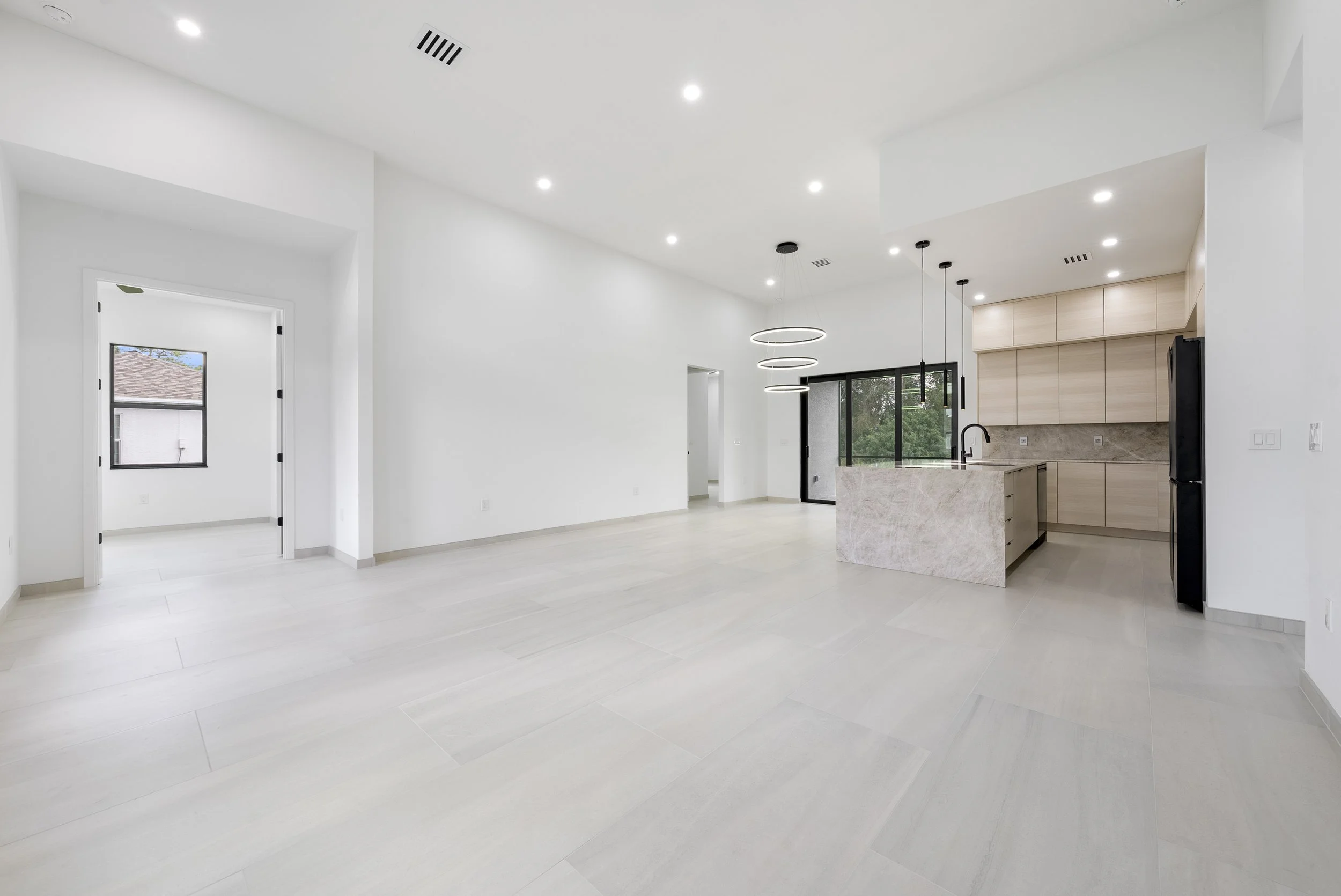 Minimalist white open-concept living space with light wood kitchen cabinets, marble kitchen island, and black appliances, featuring recessed and modern ring pendant lighting.