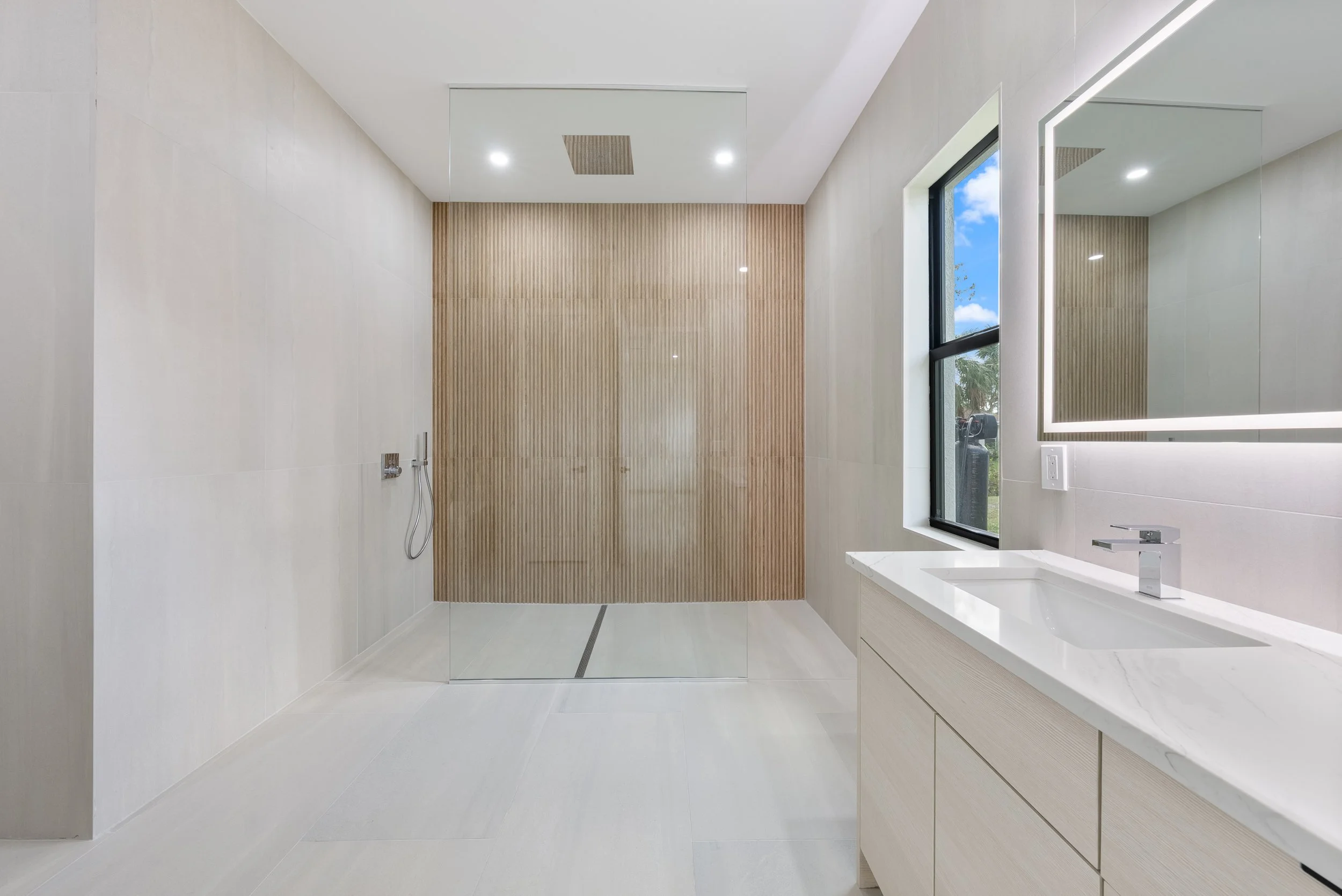 Modern bathroom with a walk-in shower with a wooden accent wall, a window, a white vanity with a marble countertop, and a large mirror with LED lighting.
