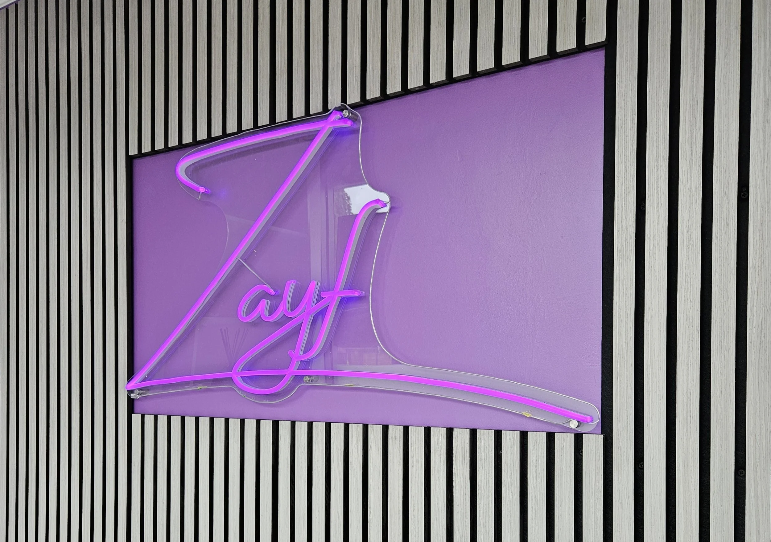Neon sign that says "Zayf" in pink and purple, mounted on a purple background with black striped wall surrounding it.