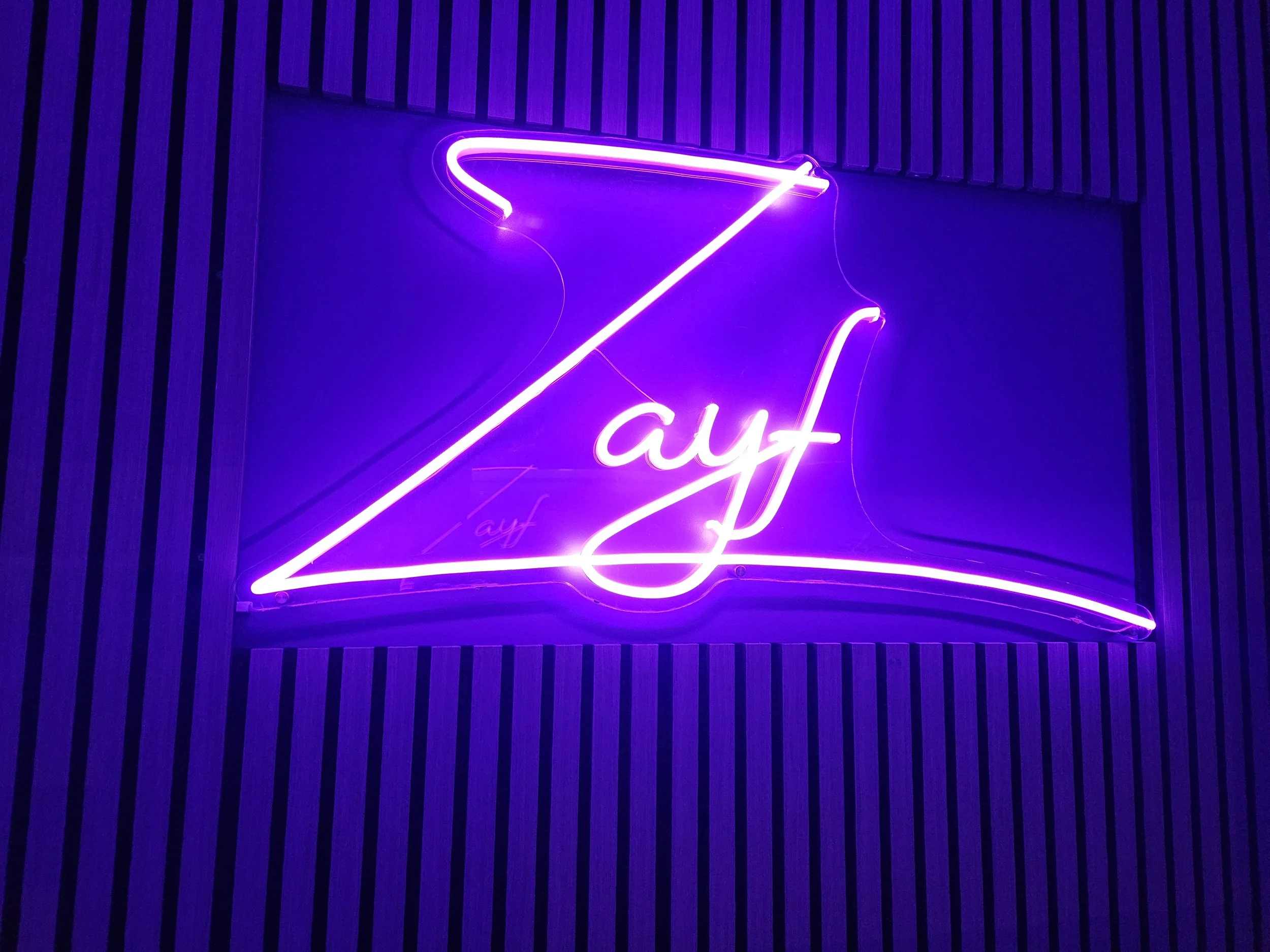 Neon sign with the word 'Zayf' in pink and purple colors on a wall with vertical black stripes.
