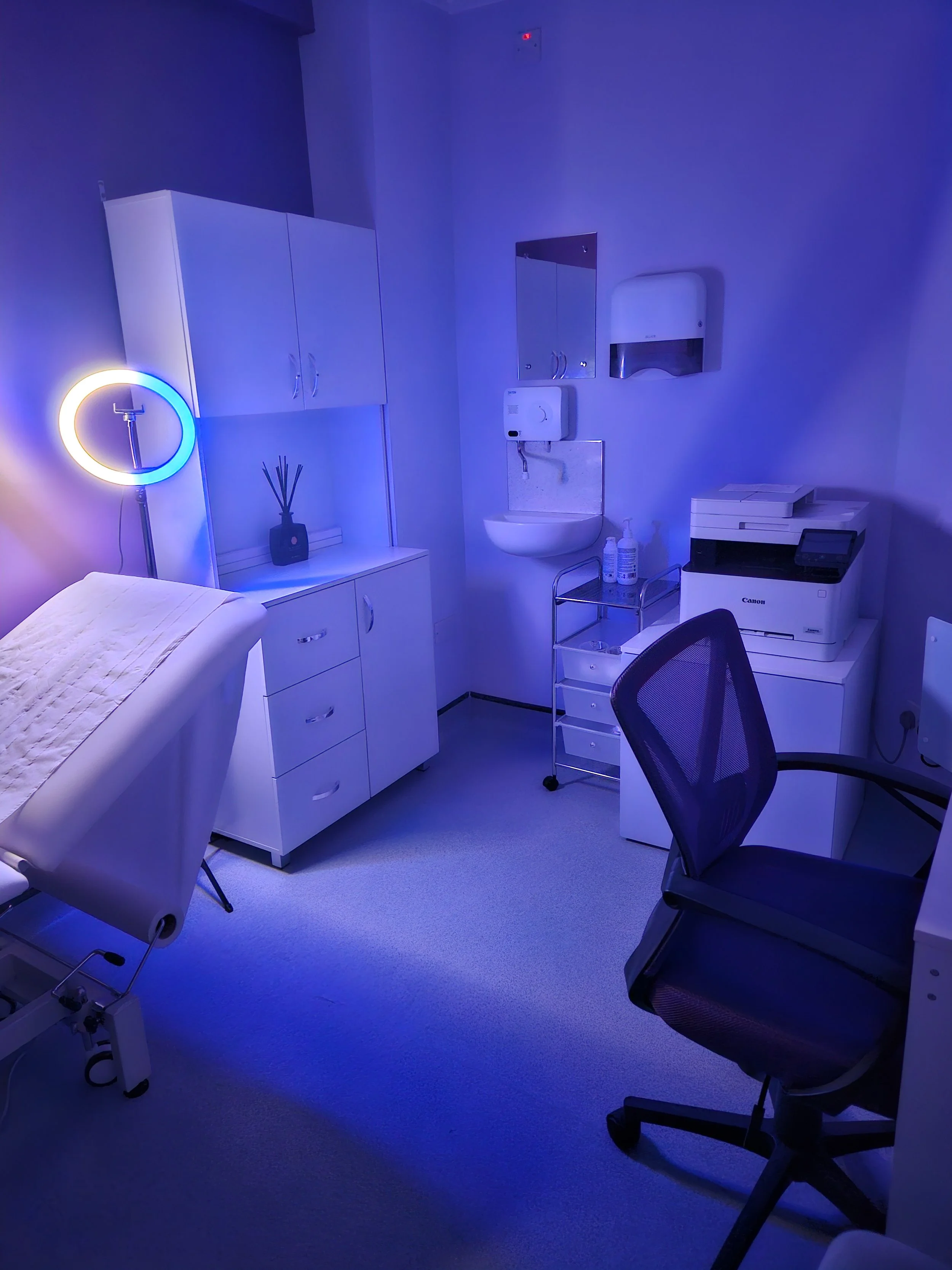 Medical examination room with a bed, a chair, cabinets, a sink, and medical equipment, illuminated with blue lighting.