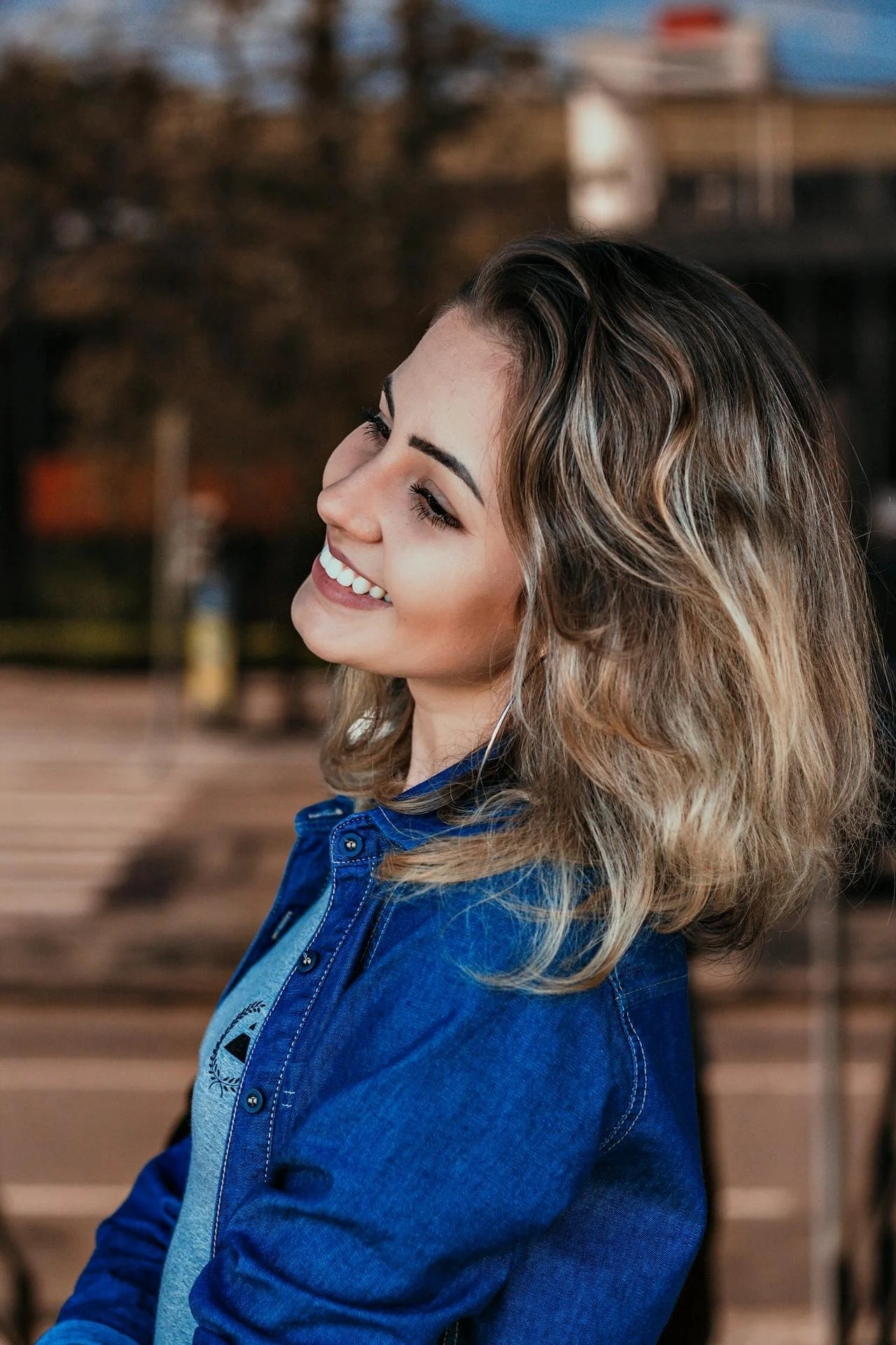 A young woman with shoulder-length wavy hair, smiling and looking to her left, wearing a blue denim jacket and a gray top, outdoors with blurred background of trees and building.
