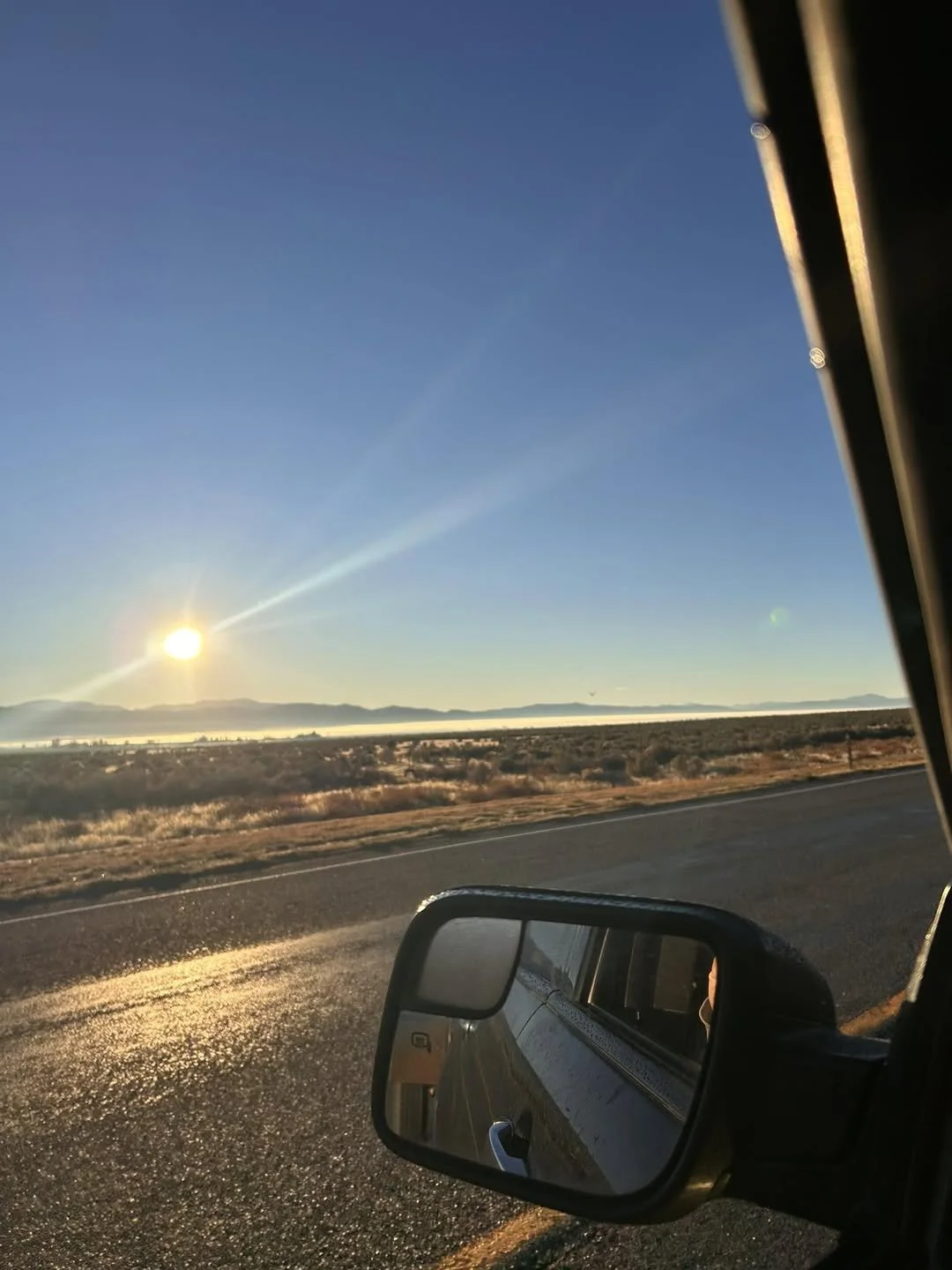 A view from inside a vehicle looking out at a rural highway at sunrise, with low mountains in the distance and the side mirror reflecting the road behind.