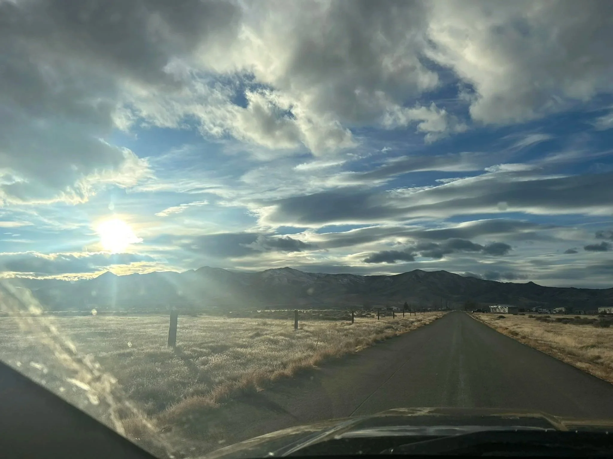 A rural road stretching toward distant mountains under a wide sky with sunlight breaking through clouds.