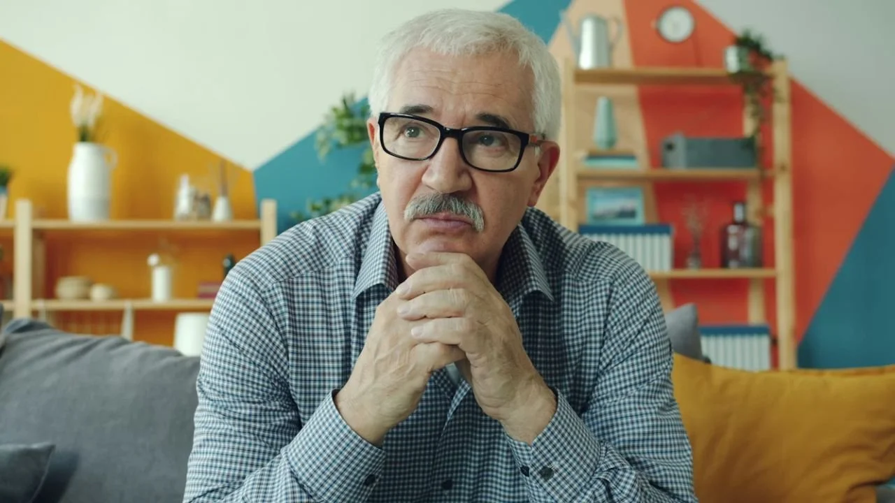 Serious senior man in glasses sitting on sofa at home thinking