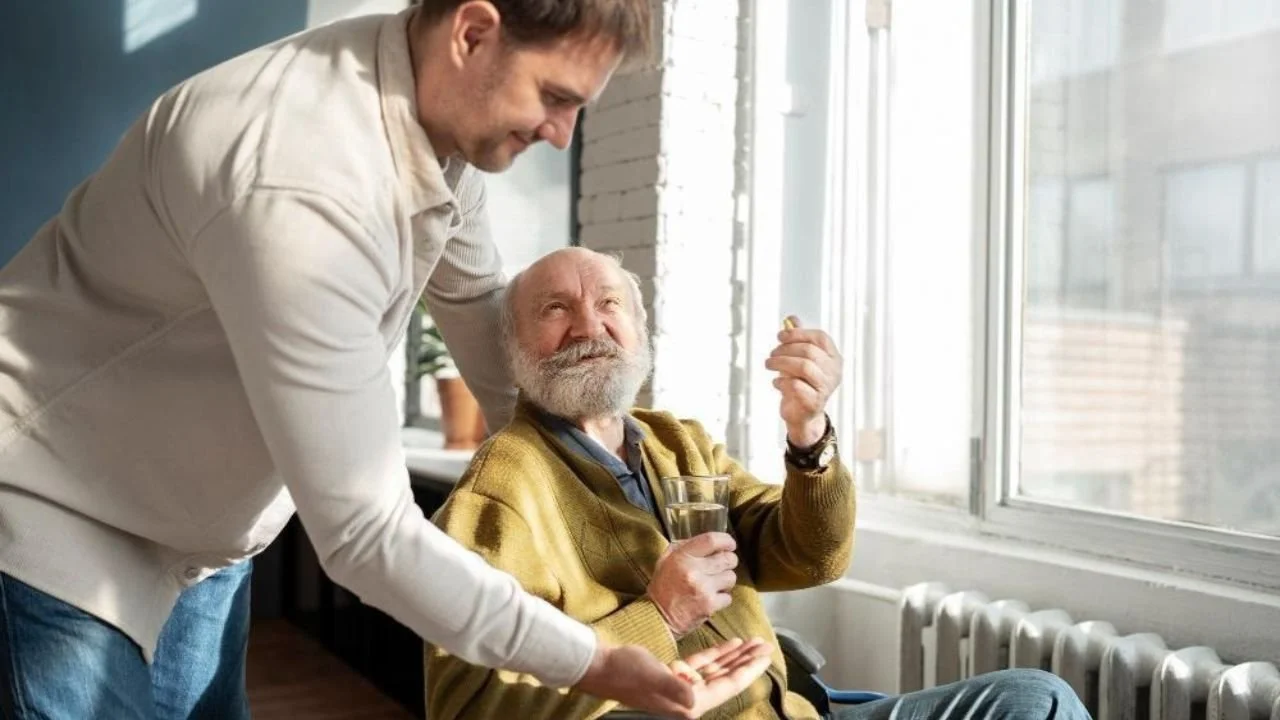 Caregiver assisting elderly man with medication near a window.