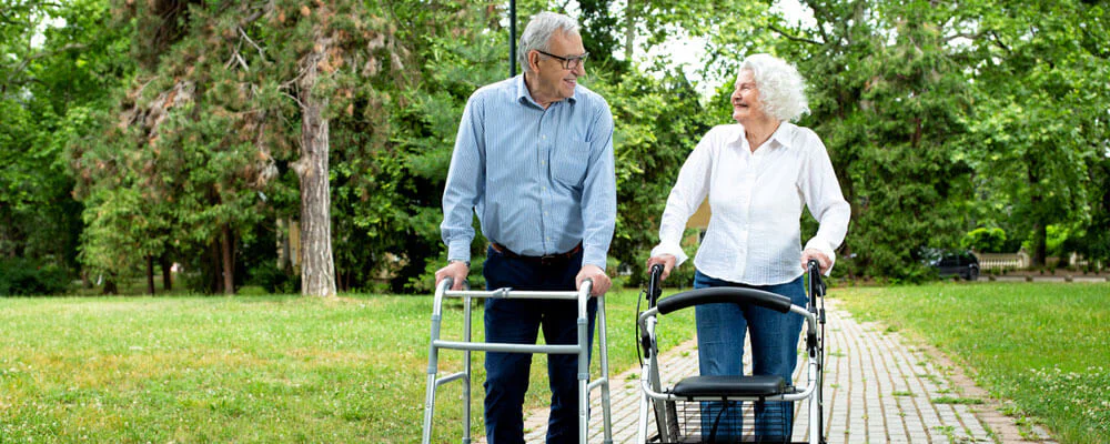 Two older adults walking with mobility aids on a green assisted living path.