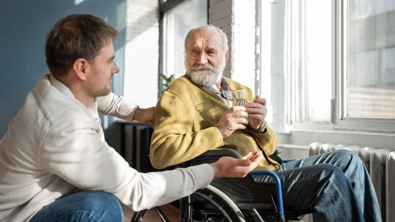 Caregiver supporting senior in wheelchair at assisted living facility window.