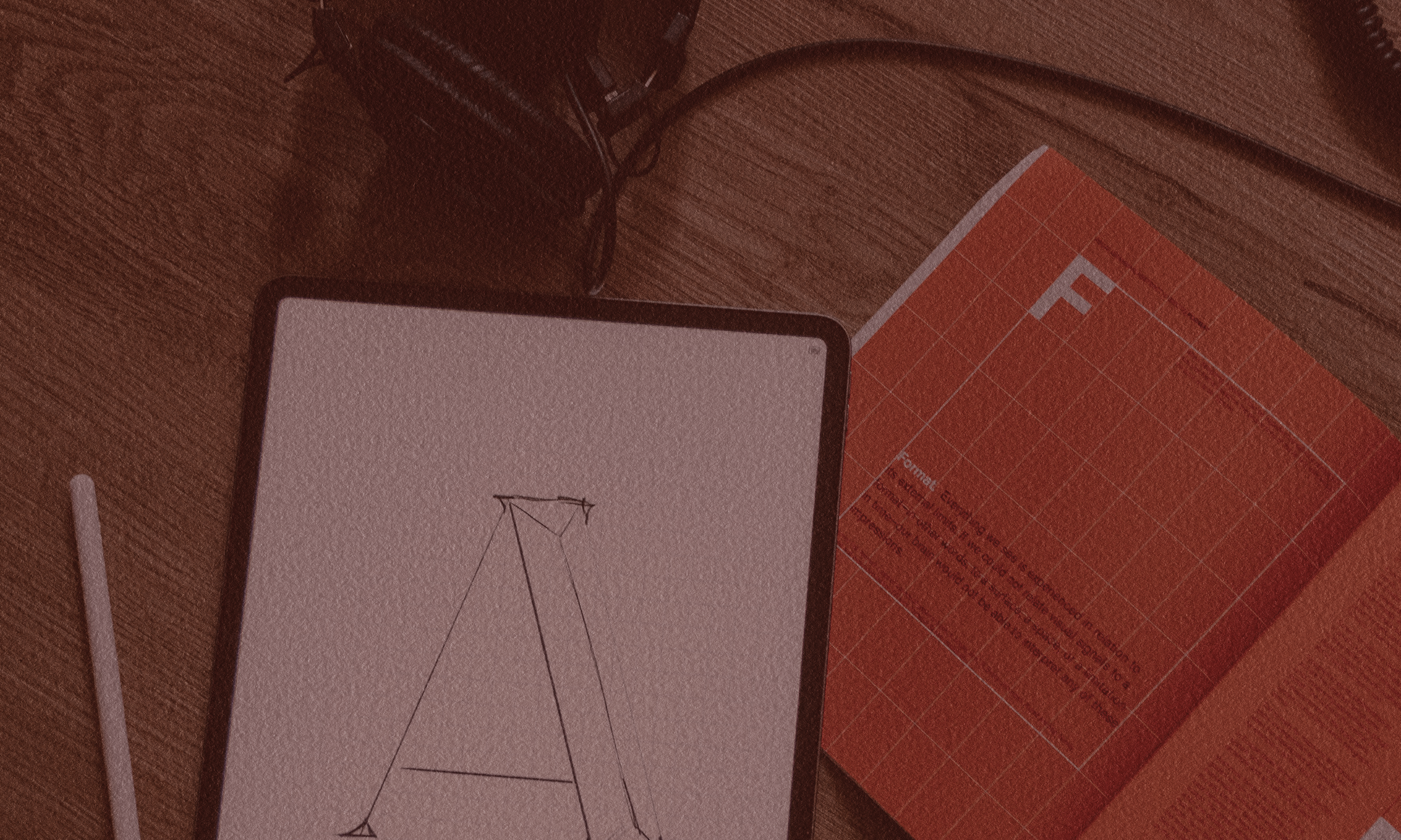 Graphic design materials such as an orange graph paper notepad and a ipad with hand drawn type in the background on a wooden surface.