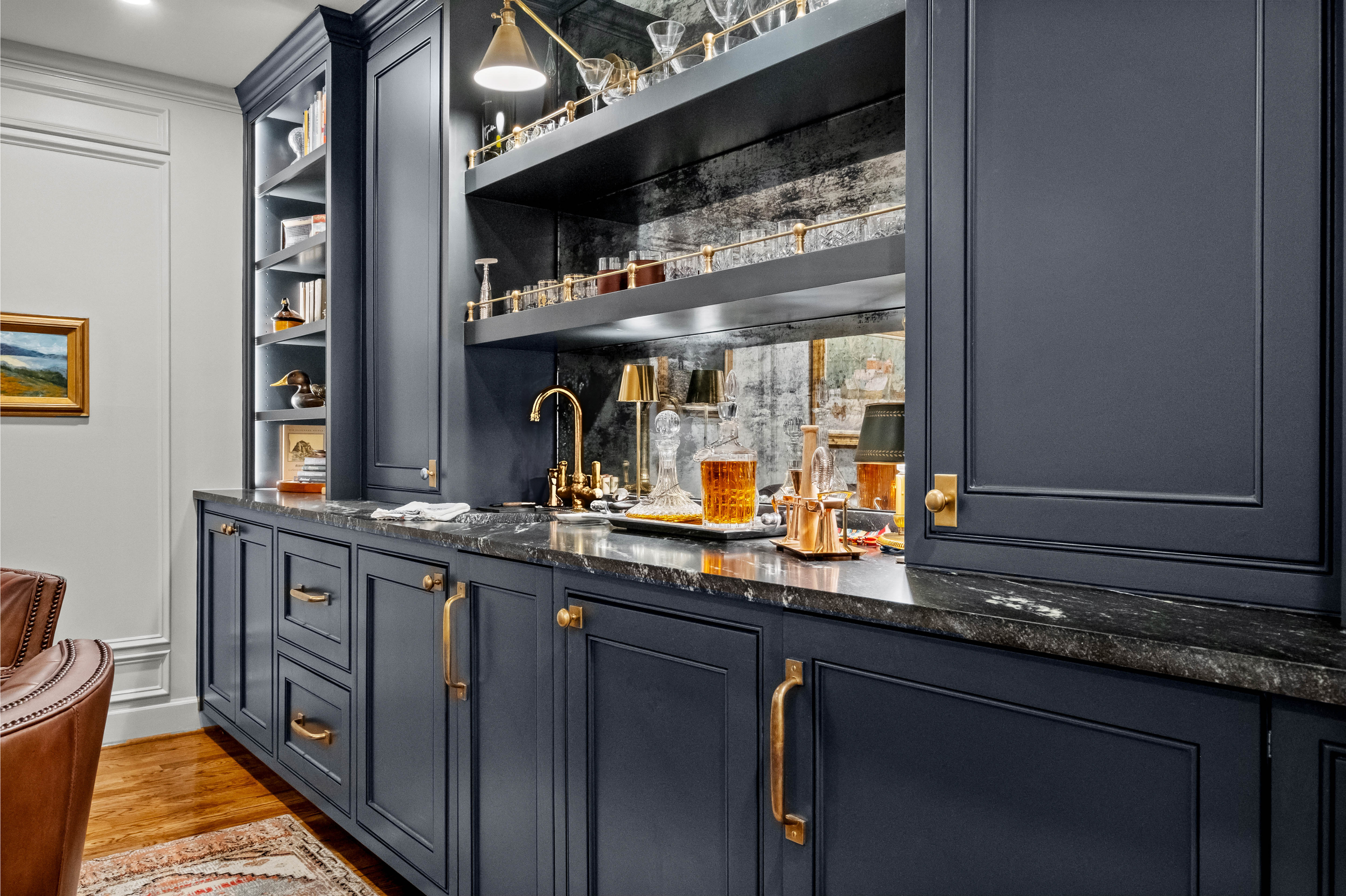 A kitchen with dark blue cabinets, gold hardware, and a black marble countertop. Glassware and decor items are on open shelves, with a window reflecting outside scenery.