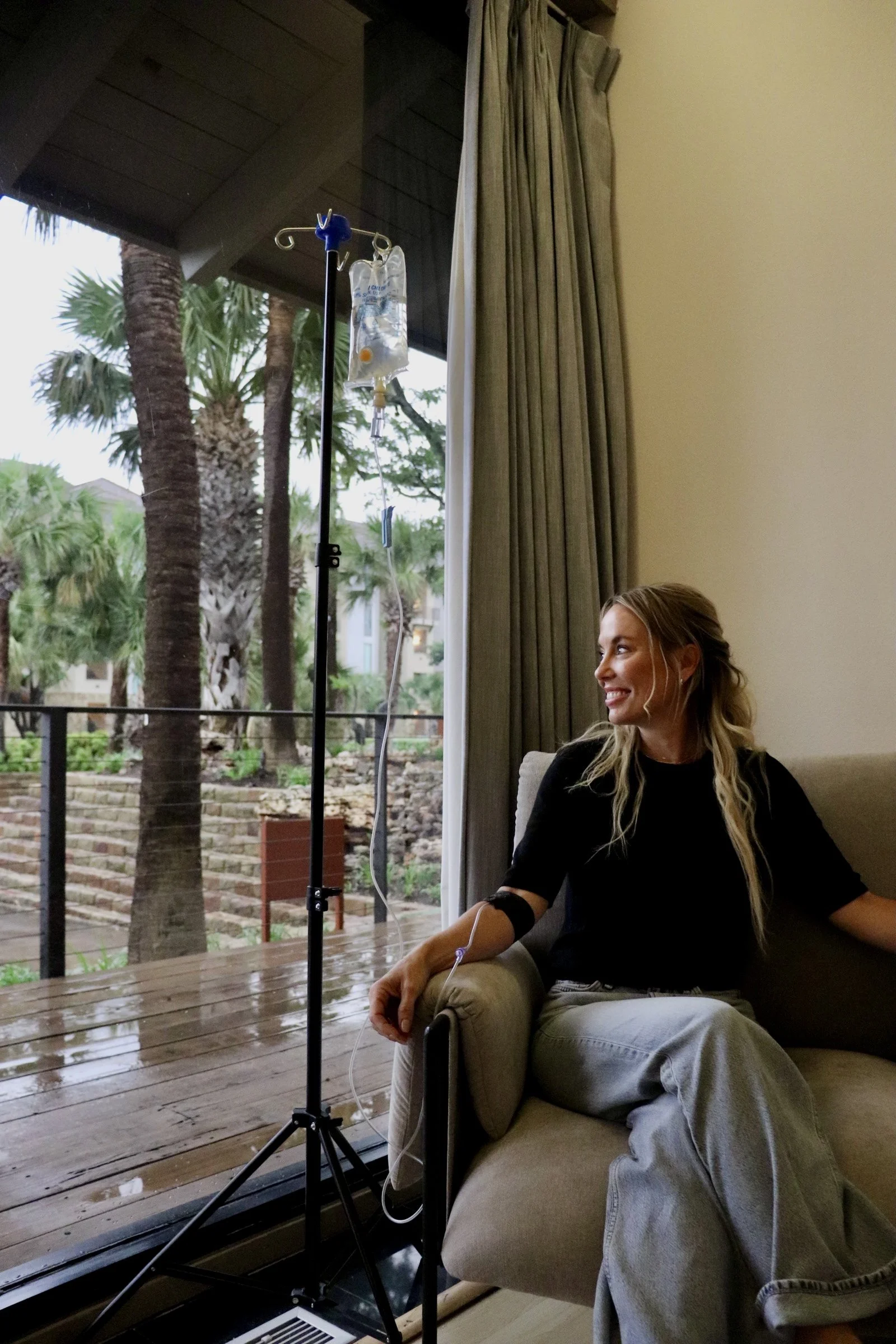 A woman sitting on a beige sofa inside a room, receiving IV therapy with a drip bag hanging from a stand, while looking outside through a sliding glass door at a garden with several palm trees.